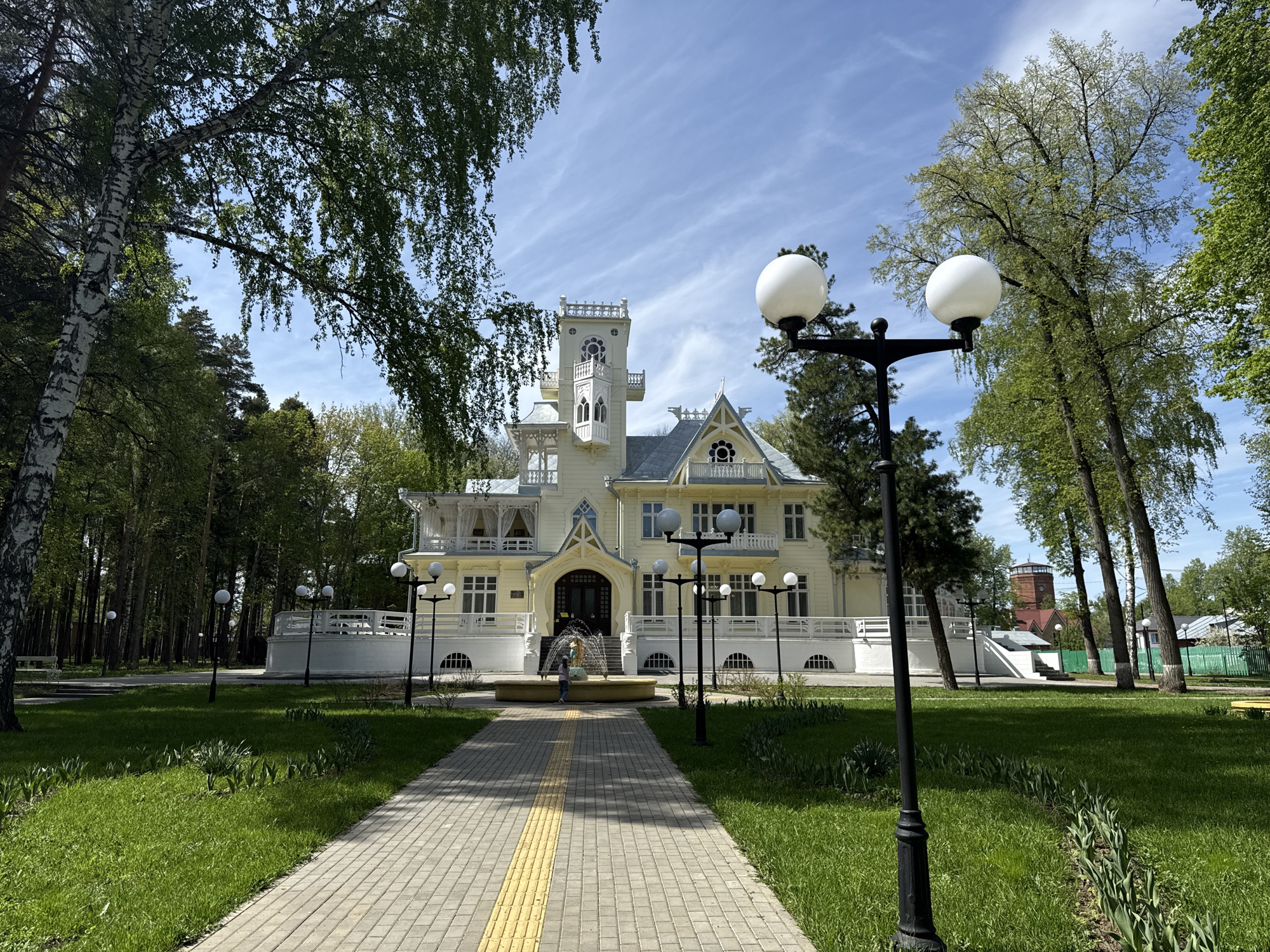 The image shows a sunny day in Sarapul, Russia, featuring a clear view of the dacha of the Bashin brothers. A stone pathway leads to the ornate, light-colored two-story building, with a central tower and decorative architectural details. A round fountain with a statue in the center stands in front of the building. A child is standing near the fountain. The pathway is lined with black lamp posts with globe lights. Green grass and trees surround the area. A tall, red brick water tower is visible in the background to the right of the building.
