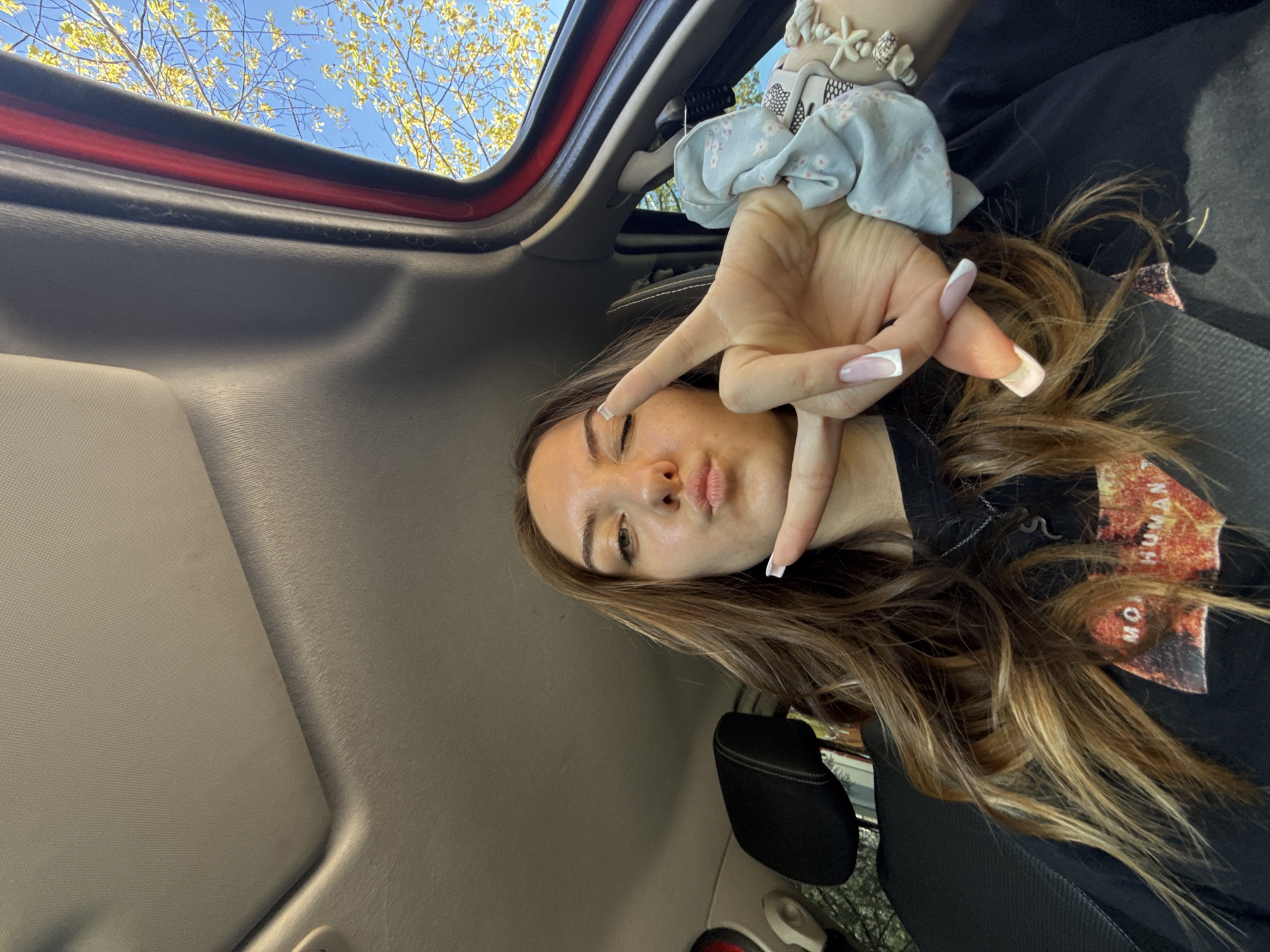 A young woman is taking a selfie inside a car. She is facing the camera with a slight pout and winking. She is making a hand gesture. She has long brown hair with blonde highlights. The interior of the car is visible, including the roof and a headrest. Outside the car, there are trees and a blue sky. The location is not discernible as Sofia, Bulgaria, from this image alone.
