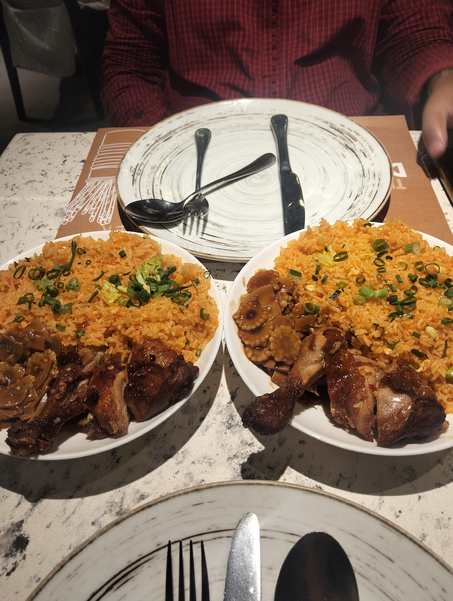 The image shows a table setting with two plates of food, likely in a restaurant in Dhaka, Bangladesh. Each plate contains fried rice garnished with green onions, accompanied by pieces of roasted chicken and what appears to be braised mushrooms. A person wearing a red plaid shirt is seated at the table. Above the food plates are two empty plates. One plate contains a spoon, fork, and knife. Another plate with knife, fork and spoon is partially shown in the lower part of the image. The plates are placed on brown placemats on a speckled beige tabletop.