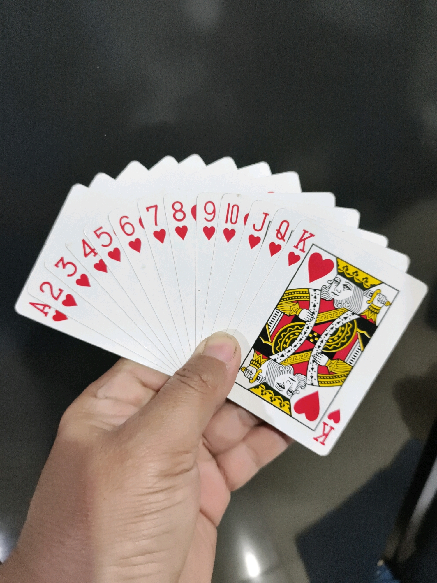 A hand is holding a fan of playing cards. The cards are all hearts and range from the 2 to the King. The King of Hearts is the furthest card to the right, and it is partially visible. The background is dark. The image was taken in Tombatu, Indonesia.
