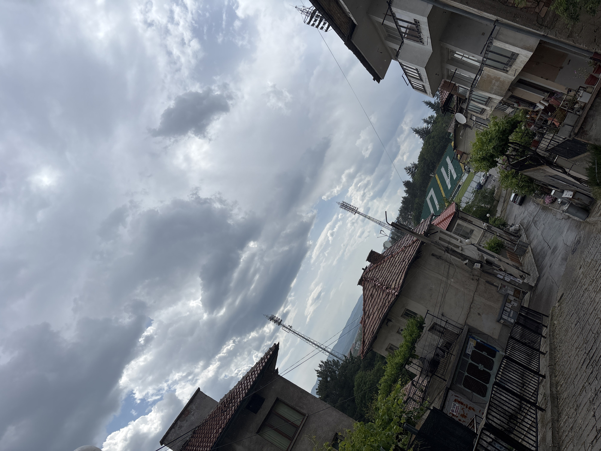 The image depicts a view within the city of Blagoevgrad, Bulgaria, characterized by a mix of residential buildings and infrastructure. The sky is overcast with dominant cloud cover. Two telecommunication towers are visible, rising above the rooftops. The buildings are constructed of concrete and have tiled roofs. A green wall is behind the houses with 'ПИИ' written on it in white letters.
A paved street and sidewalks make up the streetscape. Vegetation, including trees and vines, is interspersed throughout the scene. No people are visible.