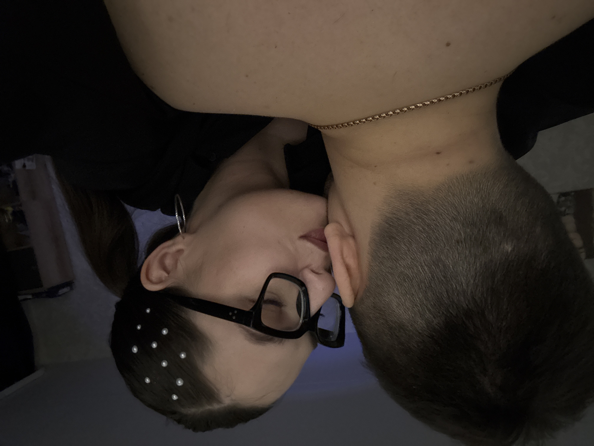 A close-up, upside-down indoor shot depicts a woman and a man in an intimate embrace. The woman has fair skin, dark hair styled in a low ponytail, black framed glasses, and small, pearl-like decorations in her hair near her hairline. She is wearing a black top and a hoop earring. She is kissing the man's neck. The man has fair skin, short, light-brown hair, and is wearing a gold chain necklace. The background is a blurry, dark gray wall. The location and city cannot be determined from this image.