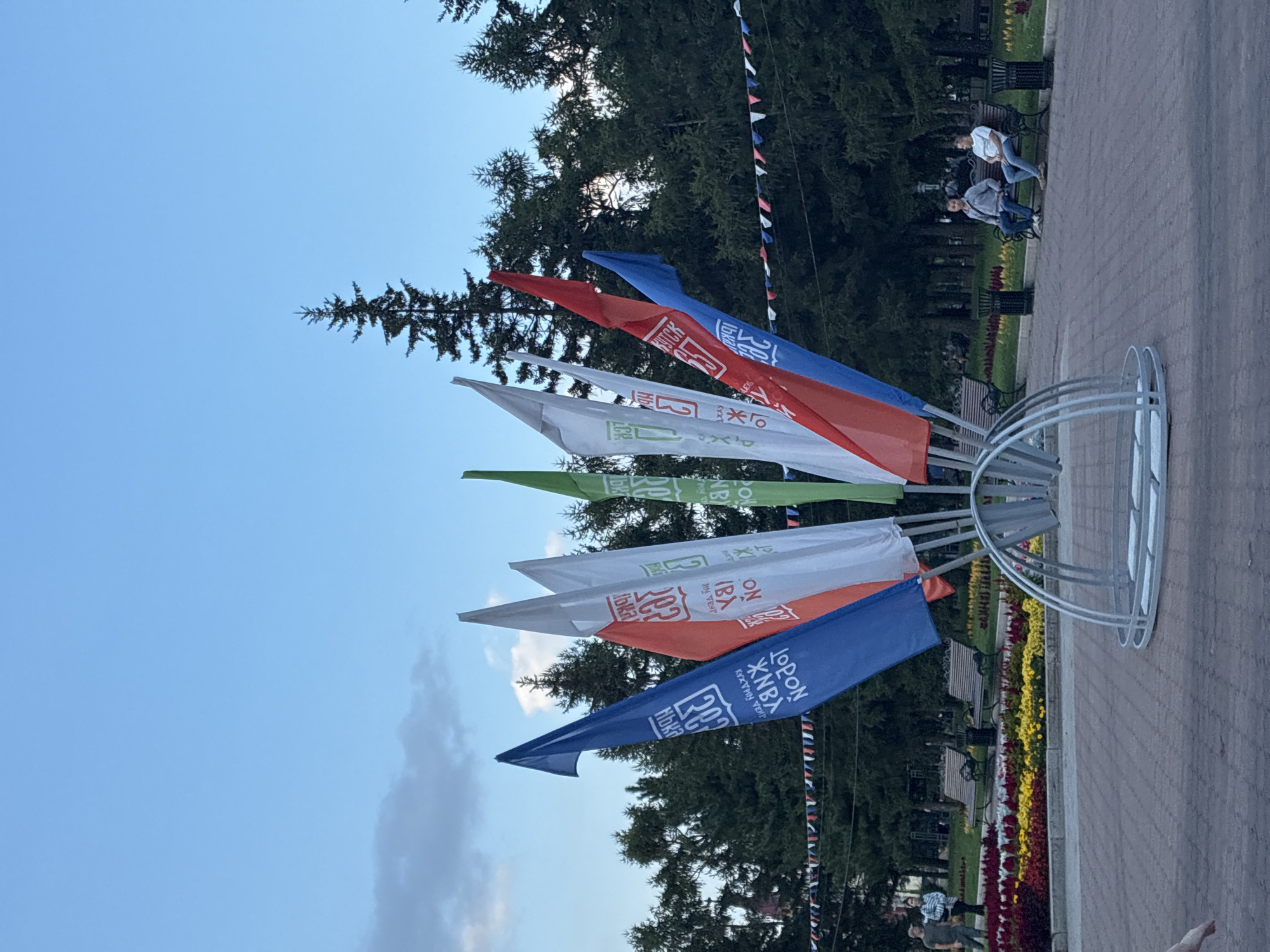 The image shows a park scene in Irkutsk, Russia, with a central display of colorful flags. The flags are arranged in a fan-like formation, with some bearing Cyrillic text and logos. In the background, there are tall evergreen trees and a bright blue sky with scattered clouds.
Several people are present in the park. Two men are sitting on a park bench in the middle ground, facing towards the right of the frame. Further in the background, near a flower bed, two individuals are walking. A decorative metal structure, possibly an art installation or part of the flag display, is prominently featured in the foreground. The ground is paved with bricks, and there are more benches and trash cans visible in the park. A string of small colorful flags is draped across some of the trees in the mid-background. The overall impression is a public park setting, possibly during a community event or festival, given the prominent flag display.