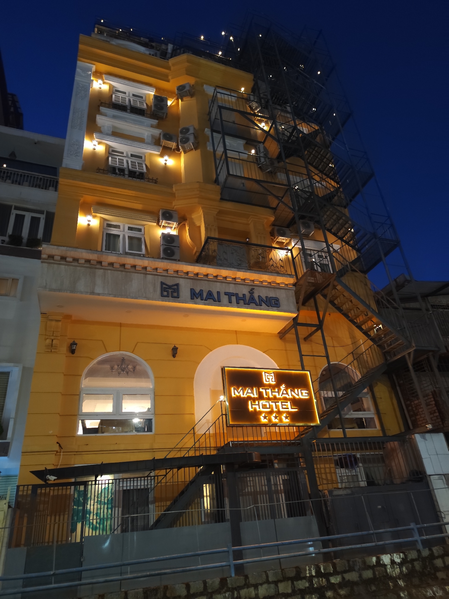 The image depicts the Mai Thang Hotel in Da Lat, Vietnam. The hotel is a multi-story building with a yellow facade. There is a fire escape on the right side of the building. The hotel's sign is illuminated and displays the hotel's name and three stars. The sky is dark blue, suggesting it is evening. There are air conditioning units visible on the exterior walls of the hotel. A fence and greenery are visible at the ground level. There are lights illuminating the building.