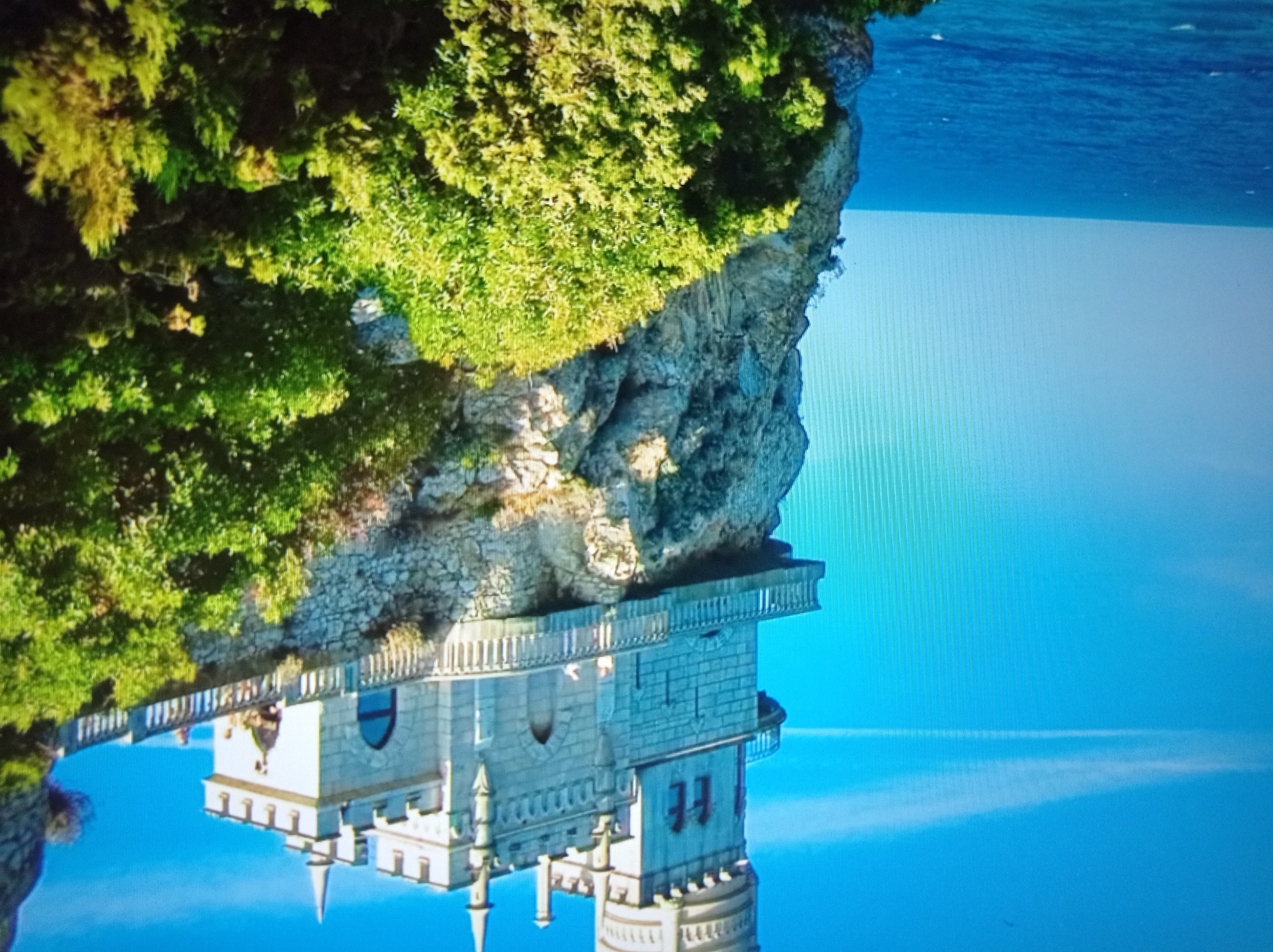 This is a photograph of the Swallow's Nest castle, a prominent landmark located on the Aurora Cliff of the Crimean Peninsula, not in Mukachevo, Ukraine. The castle, built in a neo-Gothic style, is perched dramatically on a steep cliff overlooking the Black Sea. The image captures the castle's intricate architecture, with its towers, battlements, and balconies. Lush green vegetation, including trees and shrubs, covers the cliff face and surrounds the castle, adding to its picturesque setting. The sea is a deep blue, meeting the horizon under a clear sky with a faint white cloud streak. The scene is bathed in daylight, suggesting a sunny day. There are a few indistinct figures visible on the castle's balcony.