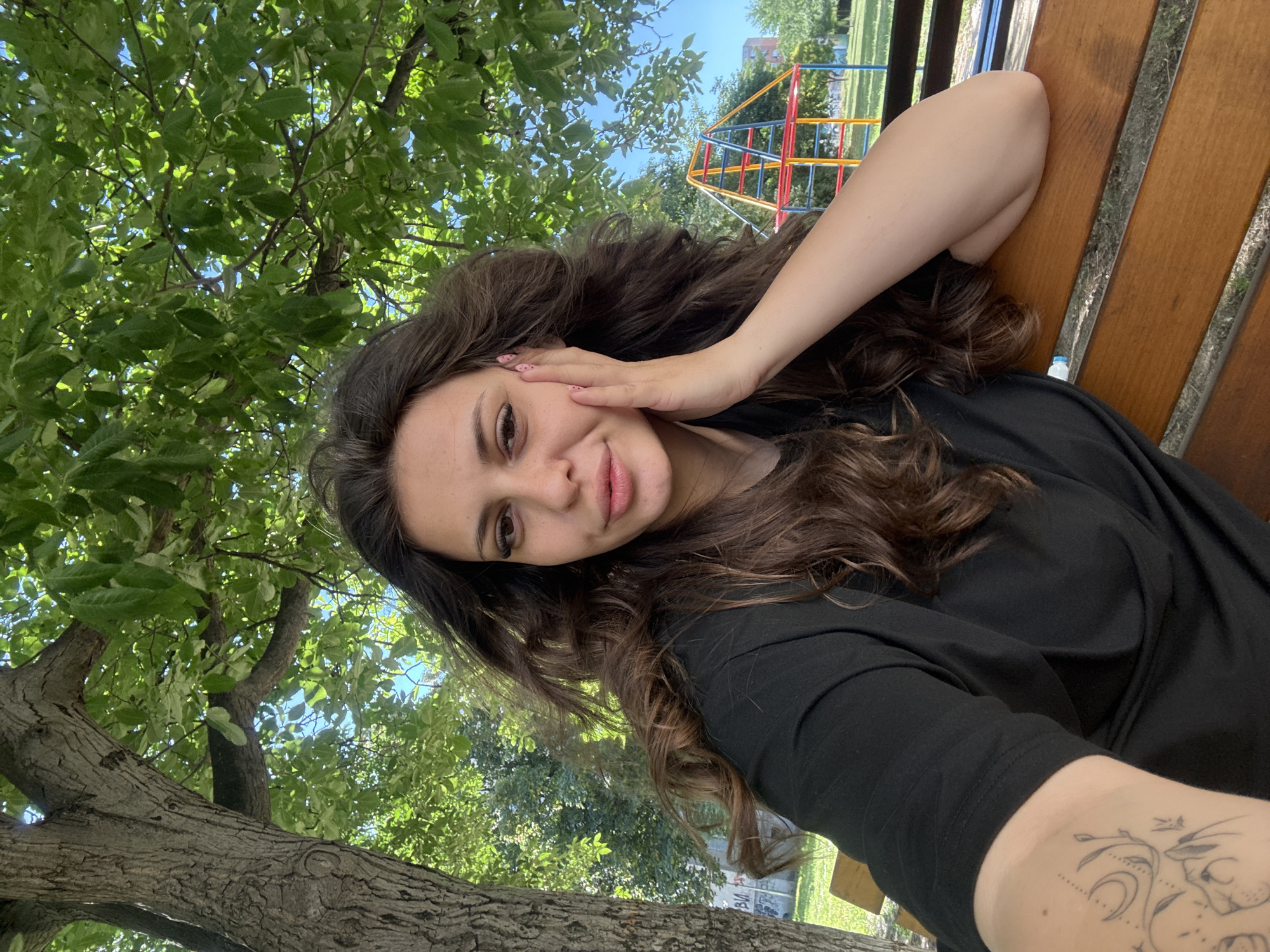 A young woman with long, dark, wavy hair is sitting on a wooden park bench, leaning her head against her hand, and looking at the camera. She is wearing a black t-shirt. Her left arm, visible in the foreground, has a tattoo of a moon and floral design. Behind her, there is a large tree with green leaves, a playground structure with red, yellow, and blue elements, and other trees and buildings in the background. The scene appears to be outdoors on a sunny day. This is likely a park in Plovdiv, Bulgaria, given the context of the request, but no specific landmarks or identifying features are present in the image to confirm this.