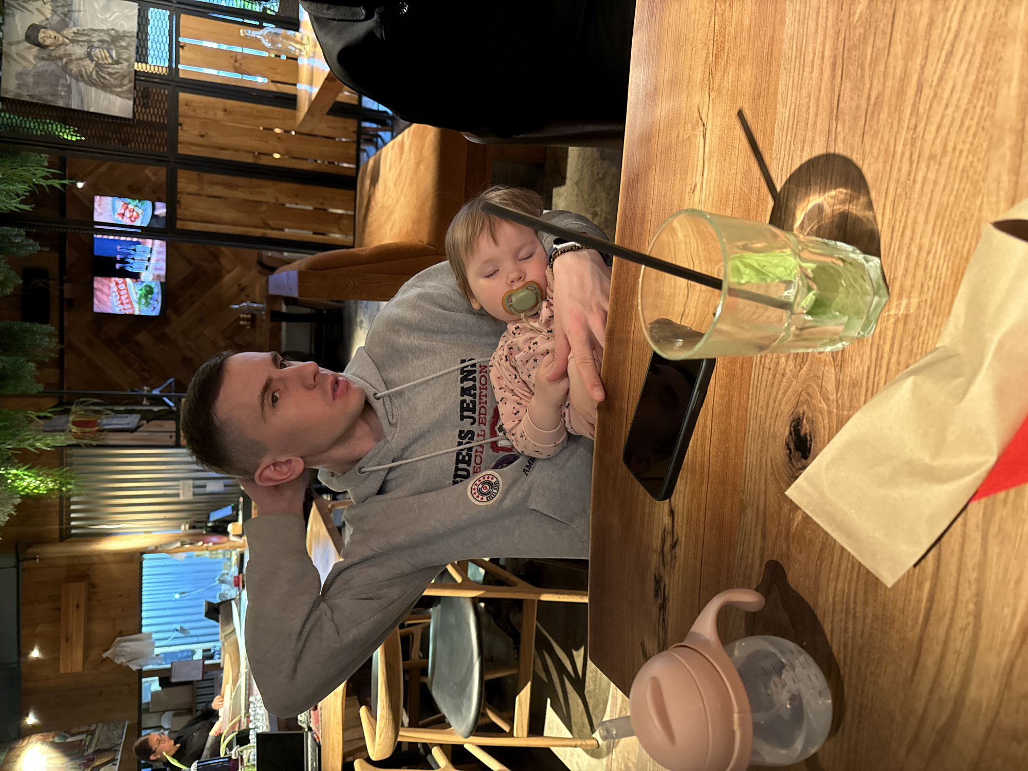A medium shot shows a man holding a sleeping baby at a table in what appears to be a restaurant. The man is light-skinned with short, dark hair. He is wearing a grey hoodie. The baby is fair-skinned with light brown hair and is sleeping with a pacifier in its mouth. The baby is wearing a pink floral patterned shirt. The table is wooden and features a drink with a straw, a smartphone, a napkin, and a baby bottle. The background shows a partially visible interior of the restaurant with wooden walls, metal structures, televisions, and other patrons. There is no mention of Malakhovka, Russia.