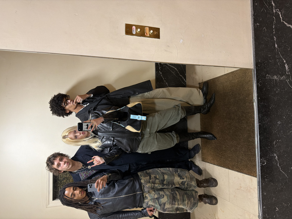 A group of four individuals pose in front of a large mirror. The first person on the left has dreadlocks. The second person is holding up the peace sign, the third person is blonde. The fourth person has an afro and eyeglasses, they have their finger up to their lip. All wear jackets. The mirror reflects a beige wall with elevator buttons, marble flooring, and a doormat. The photo is taken from a low angle and seems to be a reflection in a mirror.