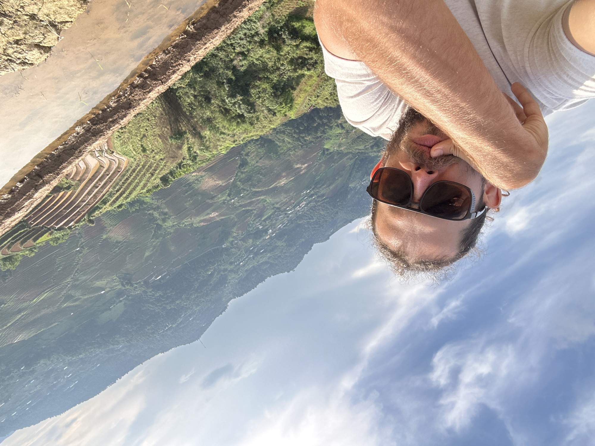 An image, initially oriented sideways, captures a man taking a selfie against a backdrop of a vast mountainous landscape. When rotated clockwise for correct viewing, a man is prominently featured in the foreground, his face partially obscured by dark sunglasses and a hand near his mouth. He wears a light-colored t-shirt and has a beard and mustache.
The background unfolds into a breathtaking panorama of extensive terraced fields, characteristic of rice paddies, stretching across lush green mountains and valleys. Some sections of the terraces appear flooded with water, reflecting the bright sky above. Small settlements or structures can be discerned within the distant valleys, nestled amidst the dense greenery. The sky is bright blue with scattered white, wispy clouds, indicating clear and sunny daytime weather over this picturesque agricultural region, consistent with the specified location of Lao Chai, Vietnam. No visible text is present in the media file.