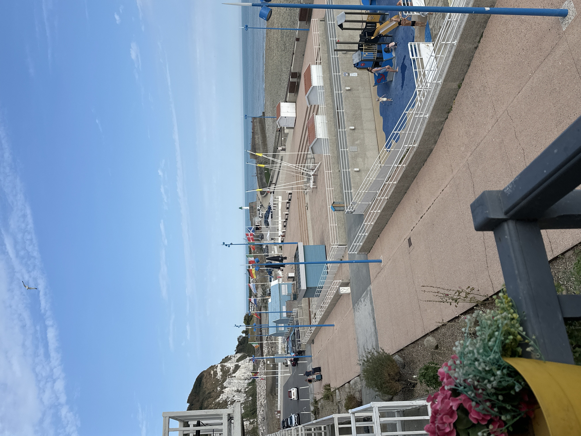 The image shows a view of the promenade and beach area in Saint-Valery-en-Caux, France, on a clear day. In the foreground, a yellow pot with pink and white flowers sits on a dark railing. The promenade, paved with reddish-brown aggregate, stretches towards the background. To the left, a steep, white-cliffed hill rises above a parking lot with several cars. A few people are walking with luggage on the promenade.
Further back, a playground with blue surfacing and various play structures, including slides and swings, is visible. Several small, white beach huts with red roofs are lined up along the promenade. Beyond the huts, a sandy beach meets the blue ocean, with a harbor wall and a lighthouse in the distance. Various national flags are displayed on flagpoles along the promenade. The sky is blue with scattered white clouds, and a seagull is flying high above.