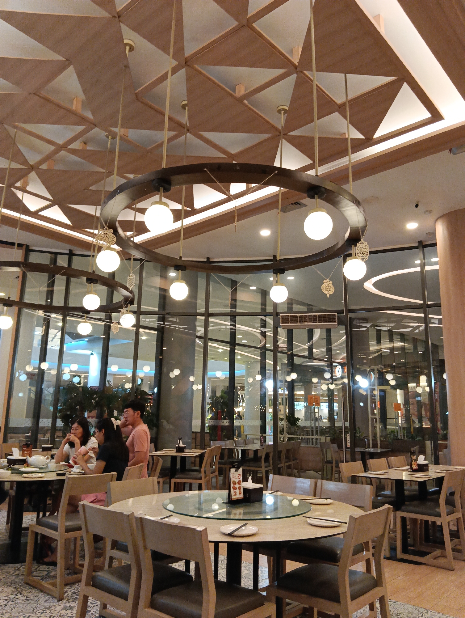 The image captures the interior of a restaurant in Sungai Raya, Indonesia, as indicated by the context provided. The scene is set within a well-lit dining area with a distinctive ceiling design composed of geometric wooden panels and white triangular inlays, illuminated by recessed lighting. Large glass windows form the exterior wall, reflecting the interior and revealing the presence of other businesses or shops in the background.

Dominating the foreground and midground are several round and rectangular dining tables with wooden chairs. The tables are set with plates, chopsticks, and teacups. In the midground, a group of three individuals, two women and one man, are seated at a round table. The man, wearing a pink t-shirt, is looking to his left, while the two women are engaged in conversation or eating. One woman, in a white top, is holding a piece of food. The other woman, wearing glasses and a black top, is also looking towards the table.

The restaurant features a large, dark circular chandelier hanging from the ceiling, from which multiple spherical pendant lights are suspended. Decorative lanterns are also hanging from the chandelier. The overall ambiance suggests a modern and somewhat upscale dining establishment. The flooring is a patterned carpet in muted tones. The reflection in the glass windows shows more tables and chairs, indicating a spacious dining area.