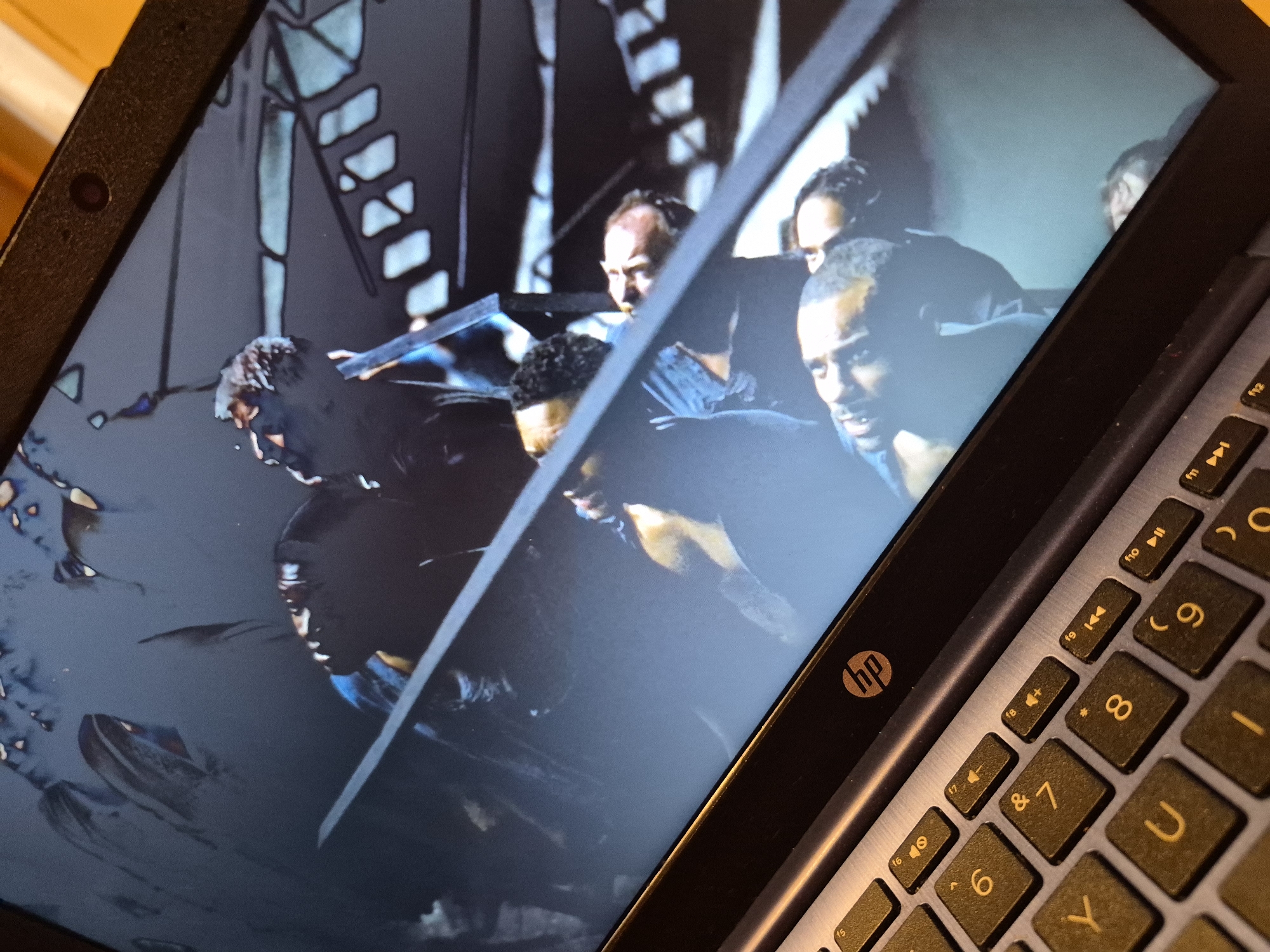 The image shows a laptop screen displaying a scene with multiple figures. The scene appears to be a still from a movie or TV show, possibly an animated or stylized production, given the distinct line-art style overlaying the figures. Several individuals are visible, some in close proximity, with dark clothing and illuminated features. The background of the screen shows an abstract, possibly metallic structure with geometric patterns. The laptop itself is silver with a black bezel, and the HP logo is visible on the top bezel. A portion of the laptop's keyboard is in the foreground, with keys labeled with numbers, letters (Y, U, I, O), and symbols (&, *). The overall lighting is dim, with a focus on the illuminated elements on the screen. There is no discernible indication of the laptop's location within Sandwell, United Kingdom.
