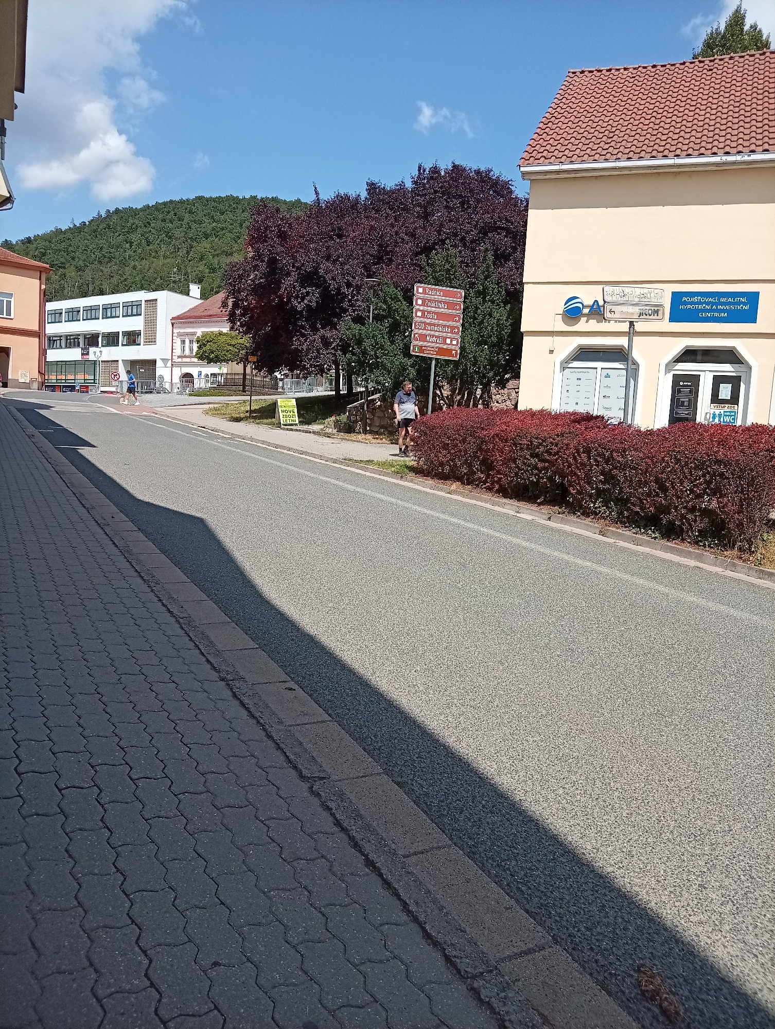 The image shows a street scene in Tišnov, Czechia, on a sunny day. In the foreground, a sidewalk made of grey interlocking pavers is visible, leading to a road with a dark grey asphalt surface. A long shadow stretches across the road from an unseen object to the left. On the right side of the road, a light yellow building with a red tiled roof houses a business. A blue sign above the entrance reads "POJIŠŤOVACÍ, REALITNI, HYPOTEČNÍ A INVESTIČNÍ CENTRUM". Next to this building is a dense, dark red bush.
Further down the street, a person in a blue shirt and shorts is riding a bicycle on the road, casting a shadow. Several street signs are visible on the left side of the road, including directional signs pointing to "Radnice", "Pošta", "SOŠ zemědělské", and "Nádraží", as well as a sign for "INFORMAČNÍ CENTRUM". Behind the signs, a large tree with dark purple foliage stands prominently.
In the background, there are several buildings, including a white building with multiple windows. Beyond the buildings, a tree-covered hillside rises under a blue sky with scattered white clouds. The overall impression is a quiet town street on a clear day.
