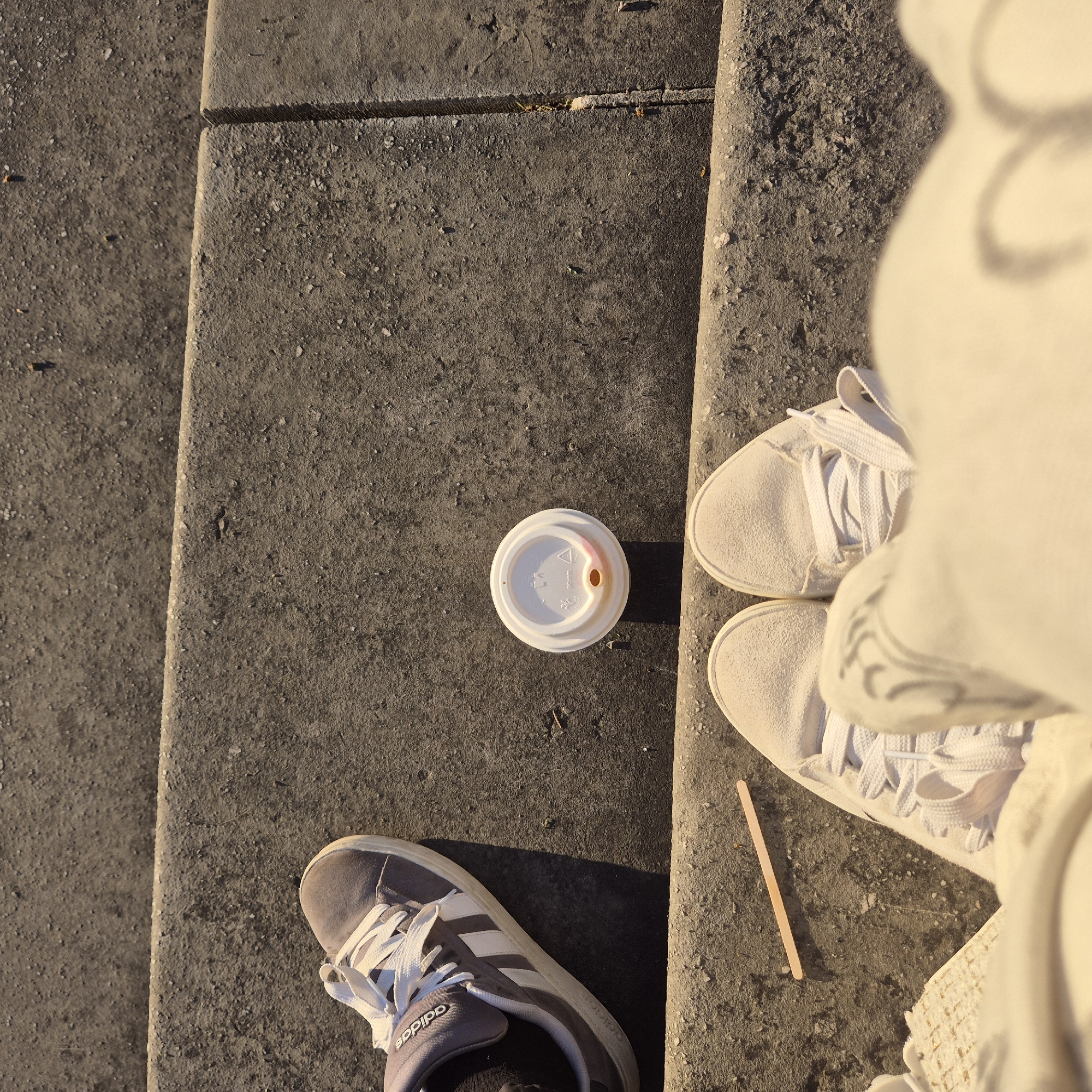 The image displays an overhead perspective of two individuals' feet on concrete steps in Strasbourg, France, under natural daylight. On the lowest visible step, a pair of feet are shod in light-colored, likely white or cream, lace-up sneakers. Adjacent to these, a light-colored wooden stirrer stick rests horizontally on the concrete. On the step immediately above and to the upper left, a single left foot is visible, wearing a grey Adidas sneaker with white stripes and white laces. A white disposable coffee cup lid, oriented face-up with a reddish-brown stain near its sip opening and visible manufacturer markings, is positioned centrally on this middle step. The concrete surfaces exhibit a coarse texture with minor particulate debris.
