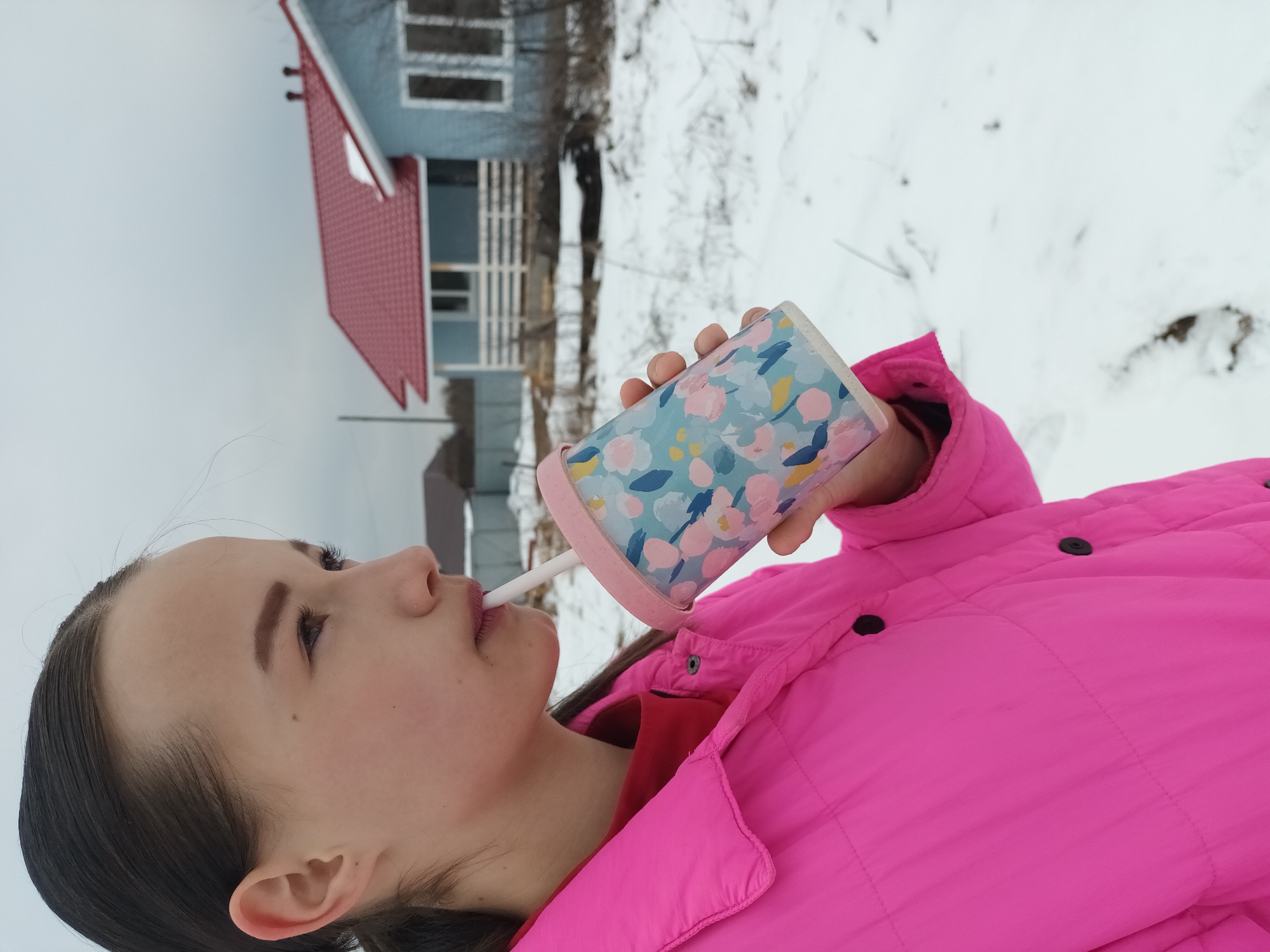 A young person drinks from a straw in Ольховка, Russia, on a snowy day. They wear a bright pink puffy jacket. The cup they are drinking from is pastel colors. A house with a red roof is in the background. The ground is covered in snow.