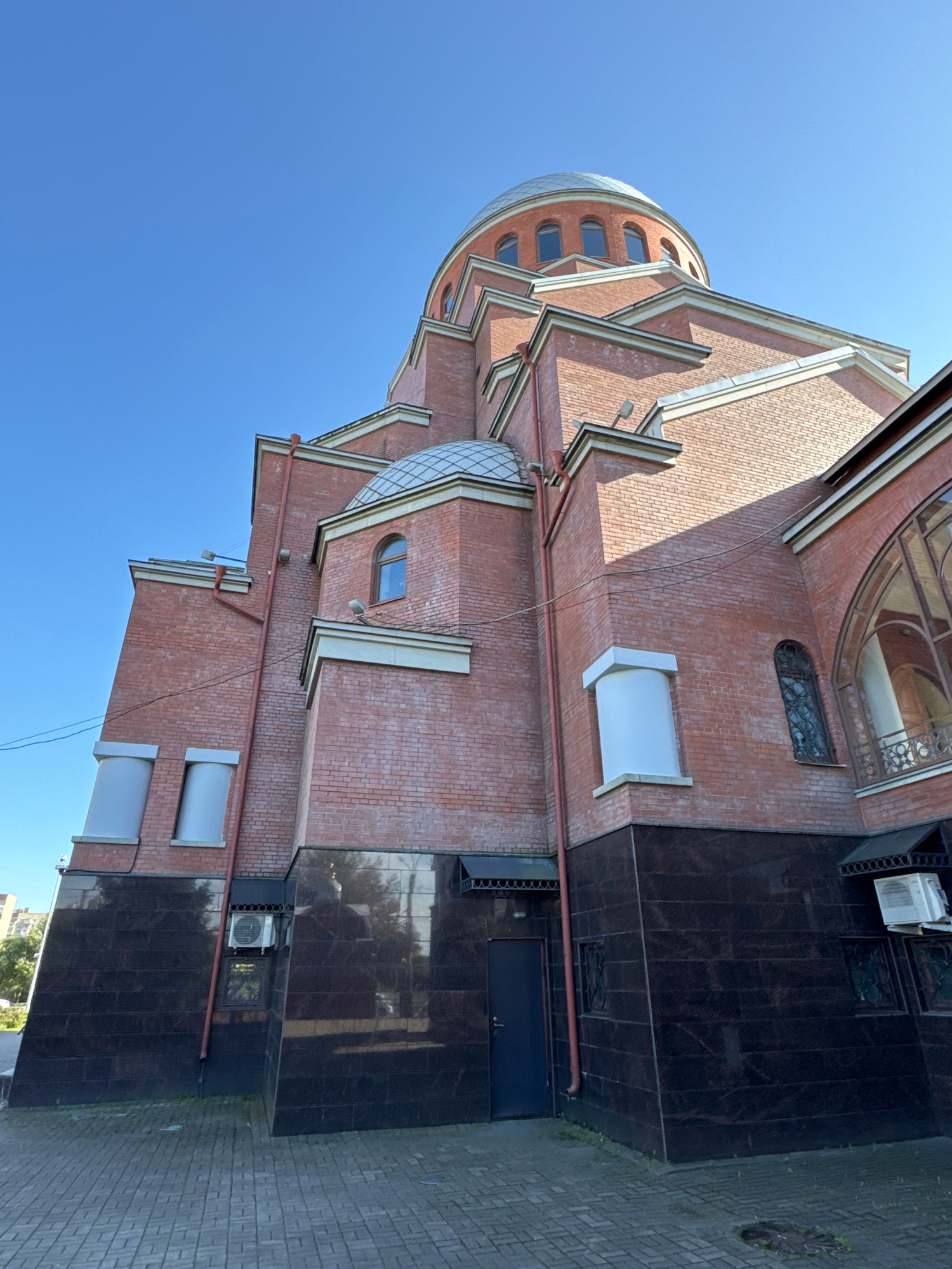 This is a low-angle, full-shot, exterior, eye-level, medium-distance photograph of the Holy Trinity Cathedral in St. Petersburg, Russia, on a clear day. The cathedral is constructed from red brick with a domed roof. The building features a dark, polished stone base, arched windows with decorative grilles, and an ornate, dark metal railing on an upper floor. There are two air conditioning units mounted on the exterior walls. The foreground is paved with grey cobblestones. The sky is a bright, clear blue. No people are visible in the image.