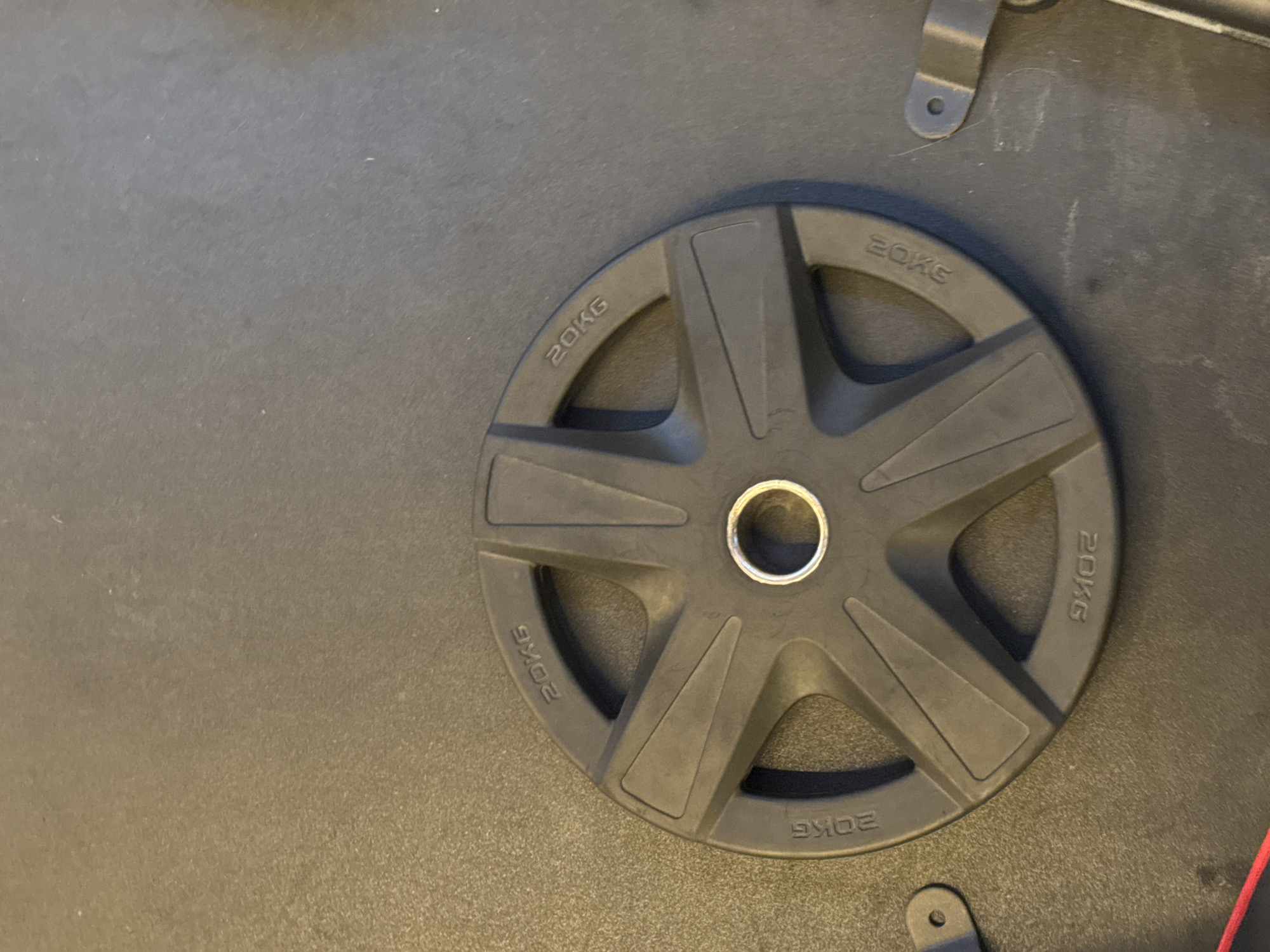 A black weight plate with a 20kg inscription is centered on a dark gray textured surface. The weight plate has five evenly spaced openings and a silver metal ring in the center. There are two small black metal clips to the left and right of the weight plate. The image appears to be taken in a gym or fitness area. The location in the city of Murcia, Spain is unknown.