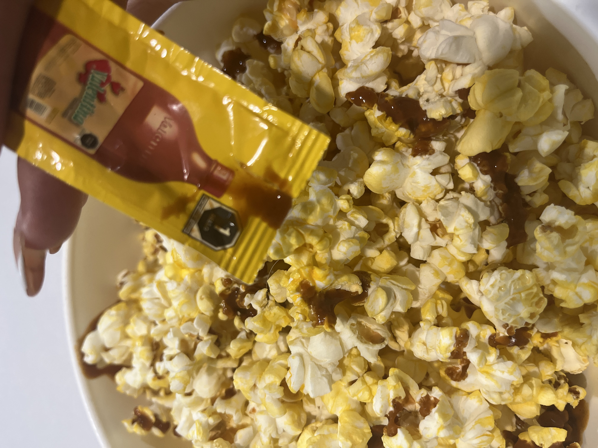 A close-up shot depicts a bowl of popcorn being drizzled with Valentina hot sauce from a yellow single-serving packet. The popcorn fills most of the frame, its kernels varying in color from pale white to buttery yellow. Streaks of dark brown Valentina sauce are visible on some of the popcorn. The packet of Valentina is held by a hand with painted fingernails. The surrounding environment is out of focus and undiscernible. There is no indication in the image where the photo was taken and there is no indication where the location of San Bernardino Tlaxcalancingo, Mexico is in the picture.