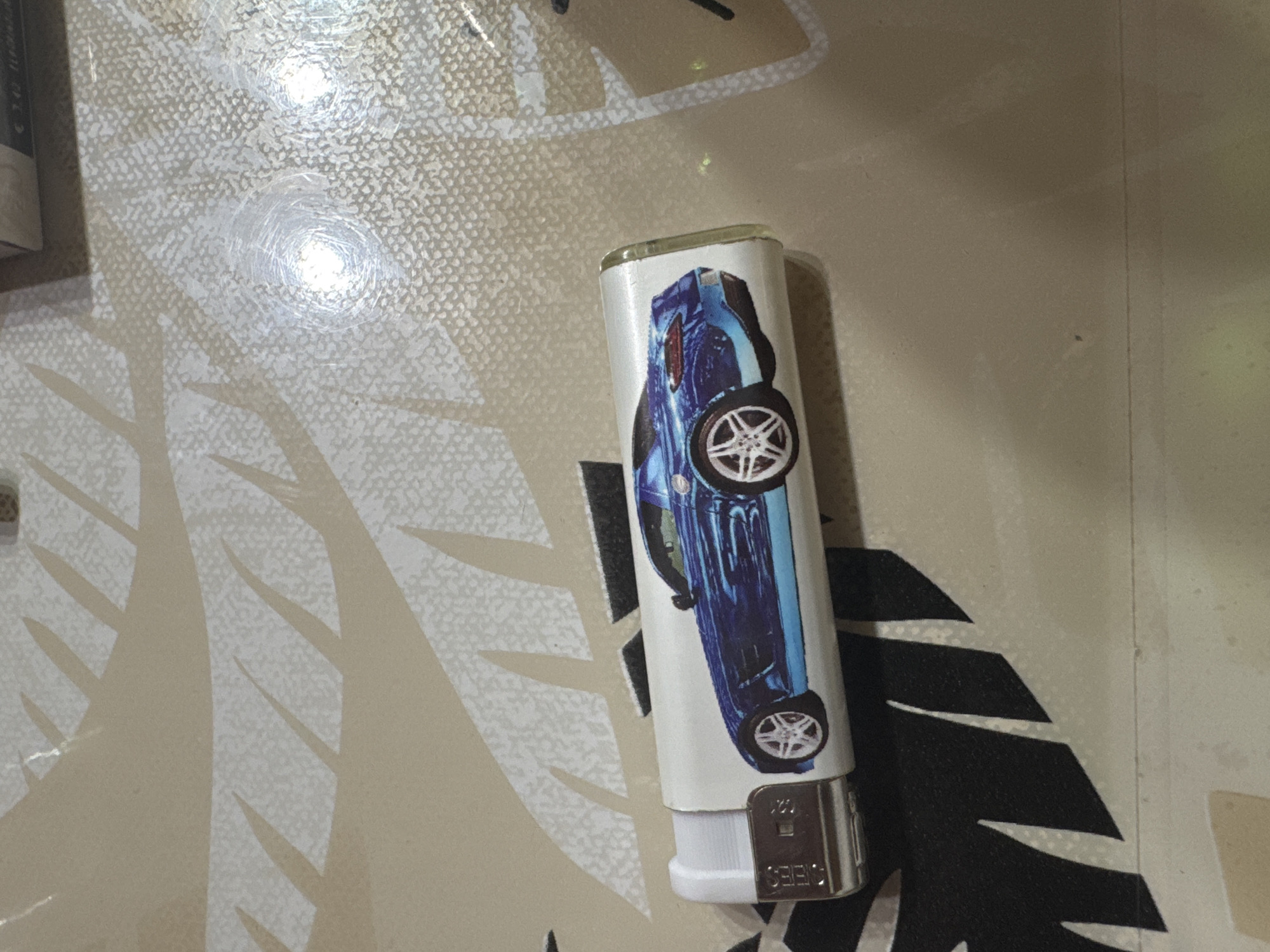 An upright, cylindrical lighter rests on a light-colored, patterned surface in Sarnevo, Bulgaria. The lighter is predominantly white, featuring a vivid blue graphic of a sleek car, possibly a sports car or coupe, shown in profile with its wheels clearly visible. The car design is oriented along the length of the lighter, with the car's front pointing towards the top. The lighter's base has a metallic silver component with visible text, including "CEBES" and "U20".
The surface beneath the lighter is a textured, light-beige material, possibly a countertop or table. It displays a repeating pattern of lighter, speckled shapes that resemble abstract foliage or organic designs, contrasting with darker, larger, angular leaf-like shapes. The surface is somewhat reflective, showing two distinct bright, circular light reflections, suggesting indoor artificial lighting. These reflections highlight numerous fine scratches and scuff marks on the surface, indicating wear. No specific activity is depicted, presenting a still life of the object.