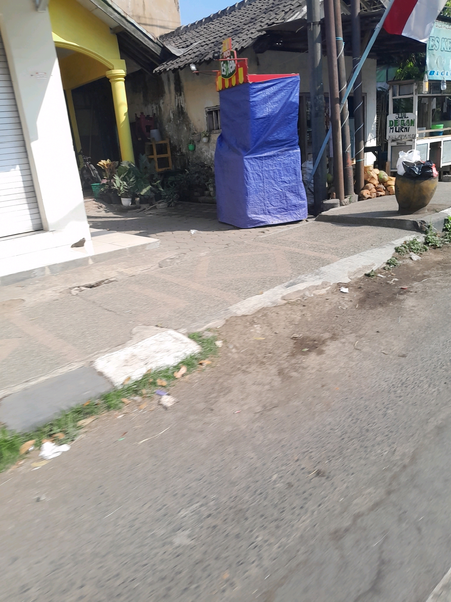 The image depicts a street scene in Ngadilangkung, Indonesia, on a sunny day. A large, blue tarp-covered structure stands on the sidewalk, possibly a temporary stall or display. To the left, a building with a yellow archway and a white rolling door is visible. Beyond the blue structure, a food stall with a sign reading "JUAL DEQAN MURNI KELAPA MUDA" is present. Coconuts are piled up near the stall, and a large clay pot filled with trash sits beside it. The street itself is paved with asphalt, and the sidewalk is made of concrete. Vegetation, including grass and potted plants, can be seen along the edge of the sidewalk and near the buildings. The overall environment suggests a commercial area with informal vending activities.