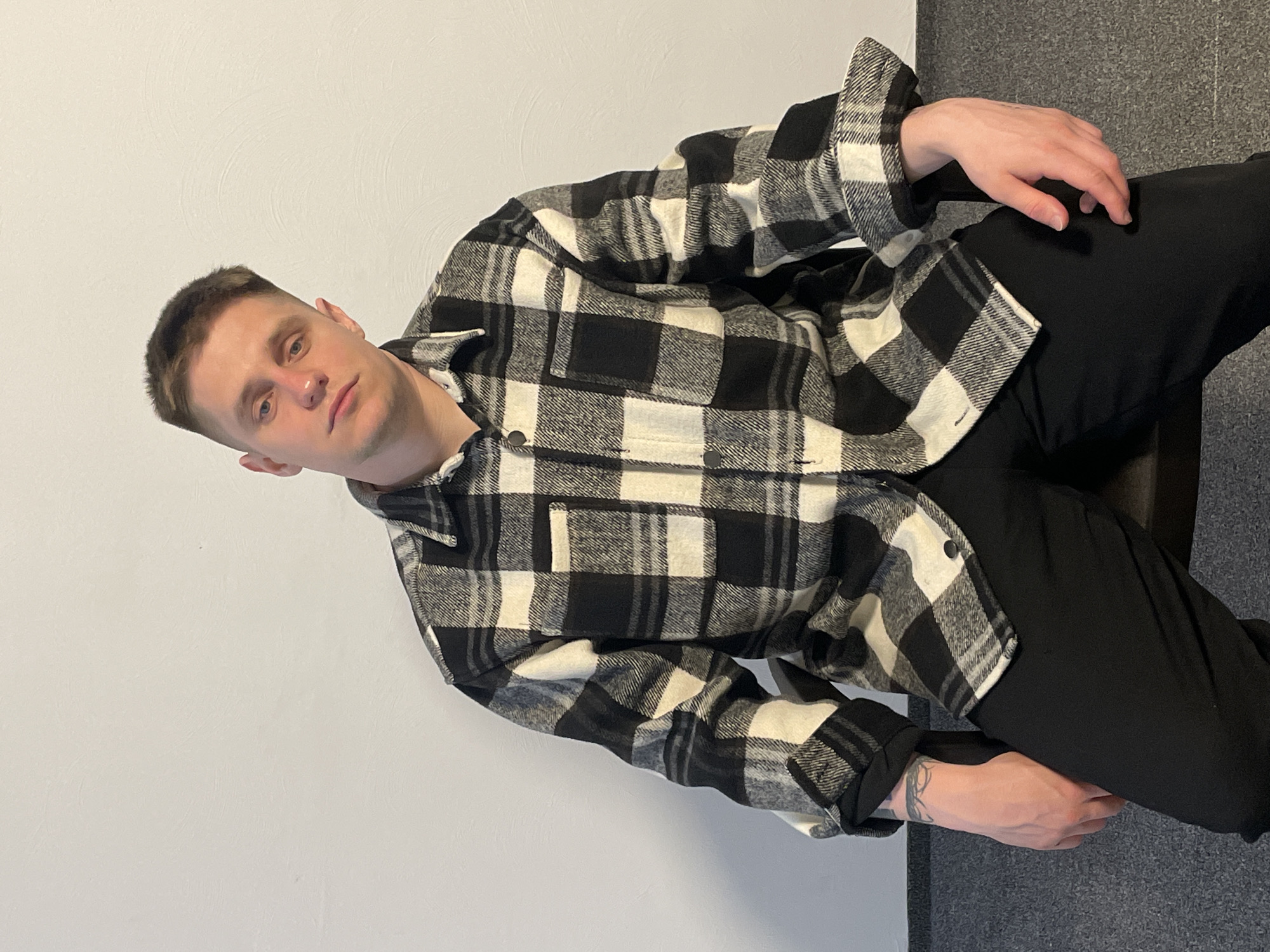 A man is sitting on a chair in front of a gray wall and a white wall. He is wearing a black and white checkered shirt and black pants. He has short brown hair and fair skin. His hands are resting on his legs. There is a tattoo on his right forearm. The location is Unknown, Unknown.