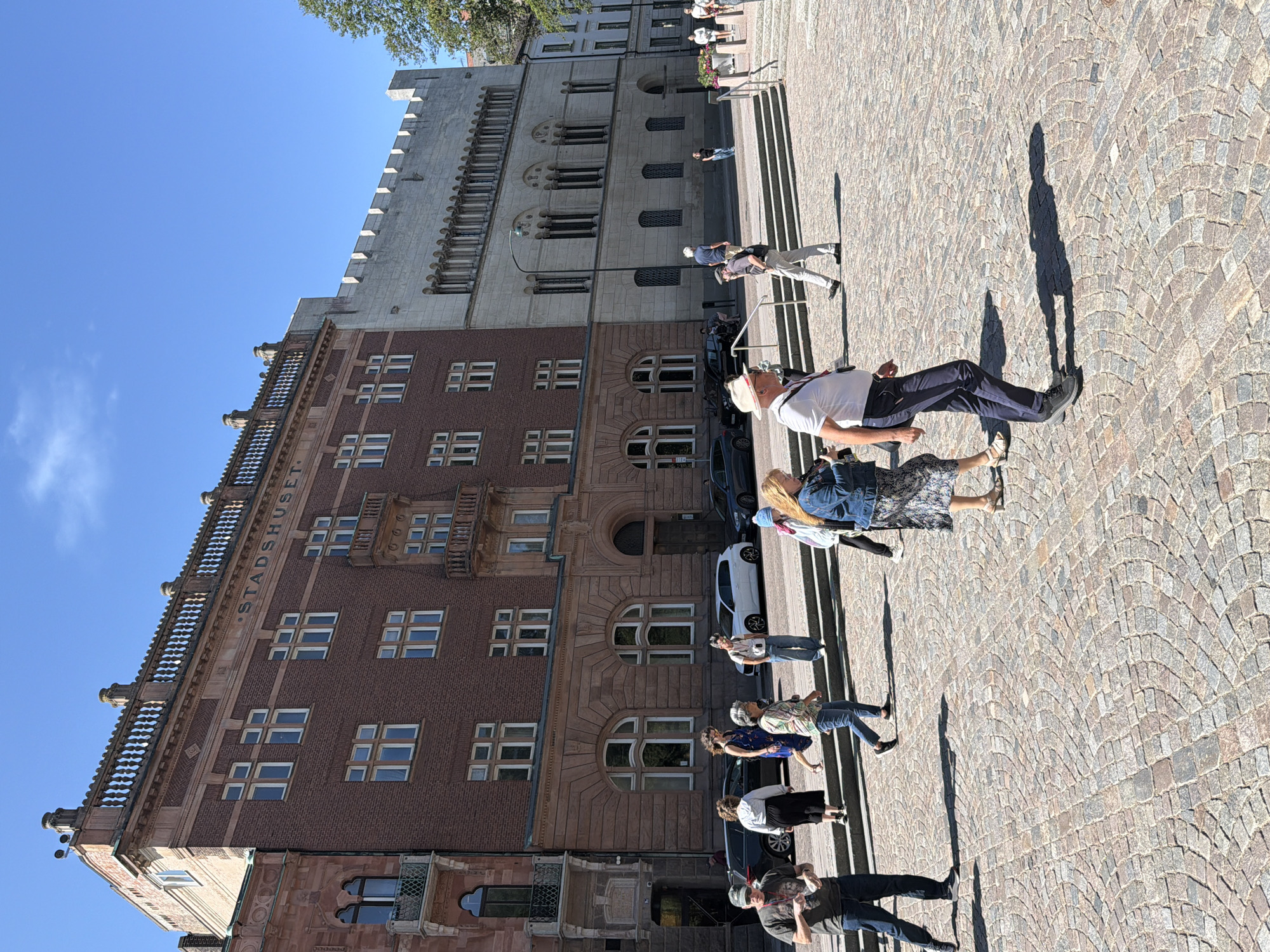 The image depicts a sunny day in Lund, Sweden, with a clear blue sky overhead. In the background, the prominent Stadshuset (City Hall) building dominates the scene. The building features a reddish-brown brick facade with intricate architectural details, including arched windows and balconies. To the right of the Stadshuset, a lighter colored building with a more modern, fortress-like appearance stands. In the foreground, a cobblestone plaza is visible, with several people walking across it. Some individuals are captured mid-stride, while others are engaged in conversation. Cars are parked along the street behind the plaza, and some foliage can be seen on the right side of the image. The overall impression is of a public square in a European city, bustling with activity on a pleasant day.