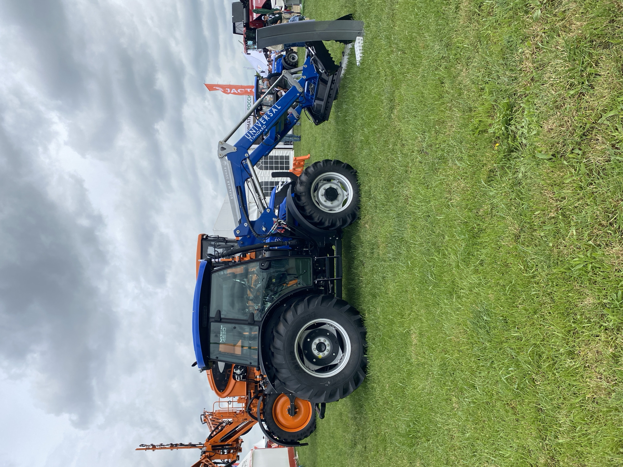 The image shows agricultural machinery on a grassy field. A blue "UNIVERSAL STANDARD" tractor with a front-end loader is prominently displayed. An orange piece of machinery is visible behind it. In the background, there is a flag with the word "JACTO" on it, along with other unidentified objects and indistinct figures. The sky is overcast. While the flag with "JACTO" on it is the most prominent feature of the image, the location in Voronezhskaya, Russia cannot be confirmed using only the given imagery. The scene depicts a possible agricultural exhibition or demonstration.
