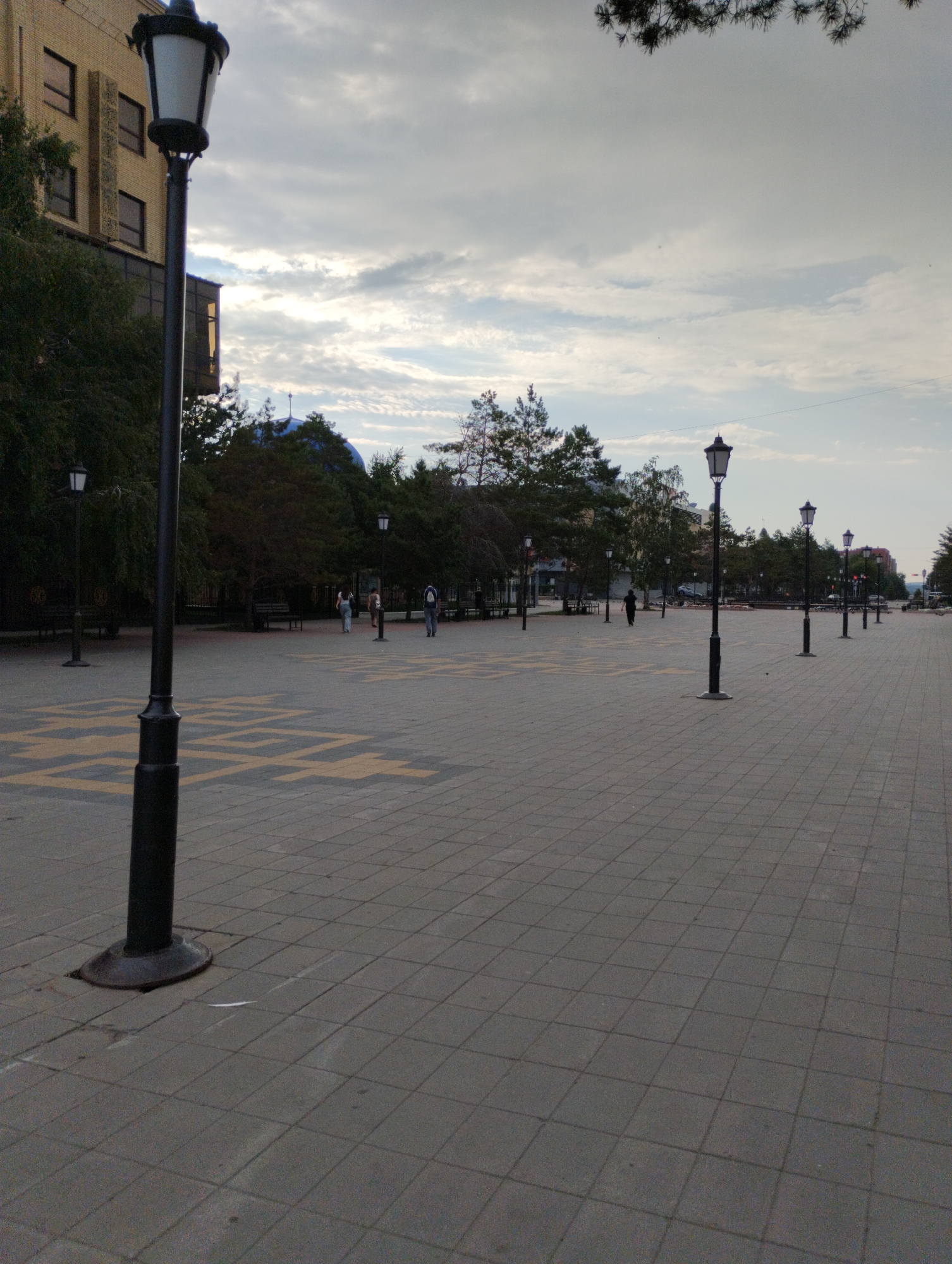 This is an outdoor, medium shot of a paved city square in Kostanay, Kazakhstan, likely on a cloudy day given the overcast sky. The foreground is dominated by a grey, tiled walkway. To the left, a dark lamppost stands prominently, its base set into the pavement. In the midground, trees line the perimeter of the square, with some people visible walking. Further in the distance, several more lampposts are arranged in a line extending into the distance. Behind the trees, buildings are visible, including a multi-story brick structure on the far left. The overall impression is one of an urban public space.