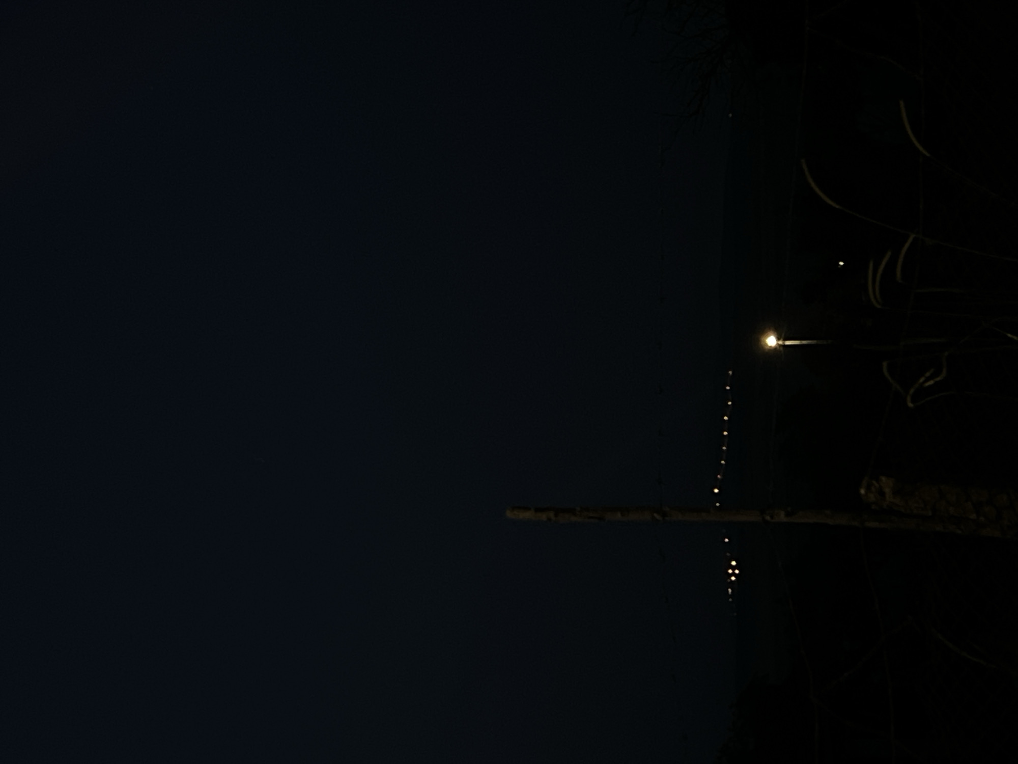 The image depicts a nighttime scene in Debelt, Bulgaria, characterized by extreme darkness and minimal artificial illumination. The upper two-thirds of the frame consist of a dark, featureless night sky. Across the mid-ground, a horizontal series of approximately twelve small, distant, yellow-orange lights suggests a remote settlement or roadway. To the left, a cluster of similar lights is also visible. In the foreground, a single, brightly glowing yellow streetlamp, positioned slightly right of the vertical center, illuminates a small area, revealing faint outlines of tall grasses or reeds to its right. To the left of the streetlamp, a dark, vertical wooden post, possibly a fence post, stands prominently. The lowest portion of the image and the areas beyond the immediate light sources are rendered in deep shadow, obscuring any detailed ground features or vegetation. There are no discernible people, animals, or vehicles in the scene.