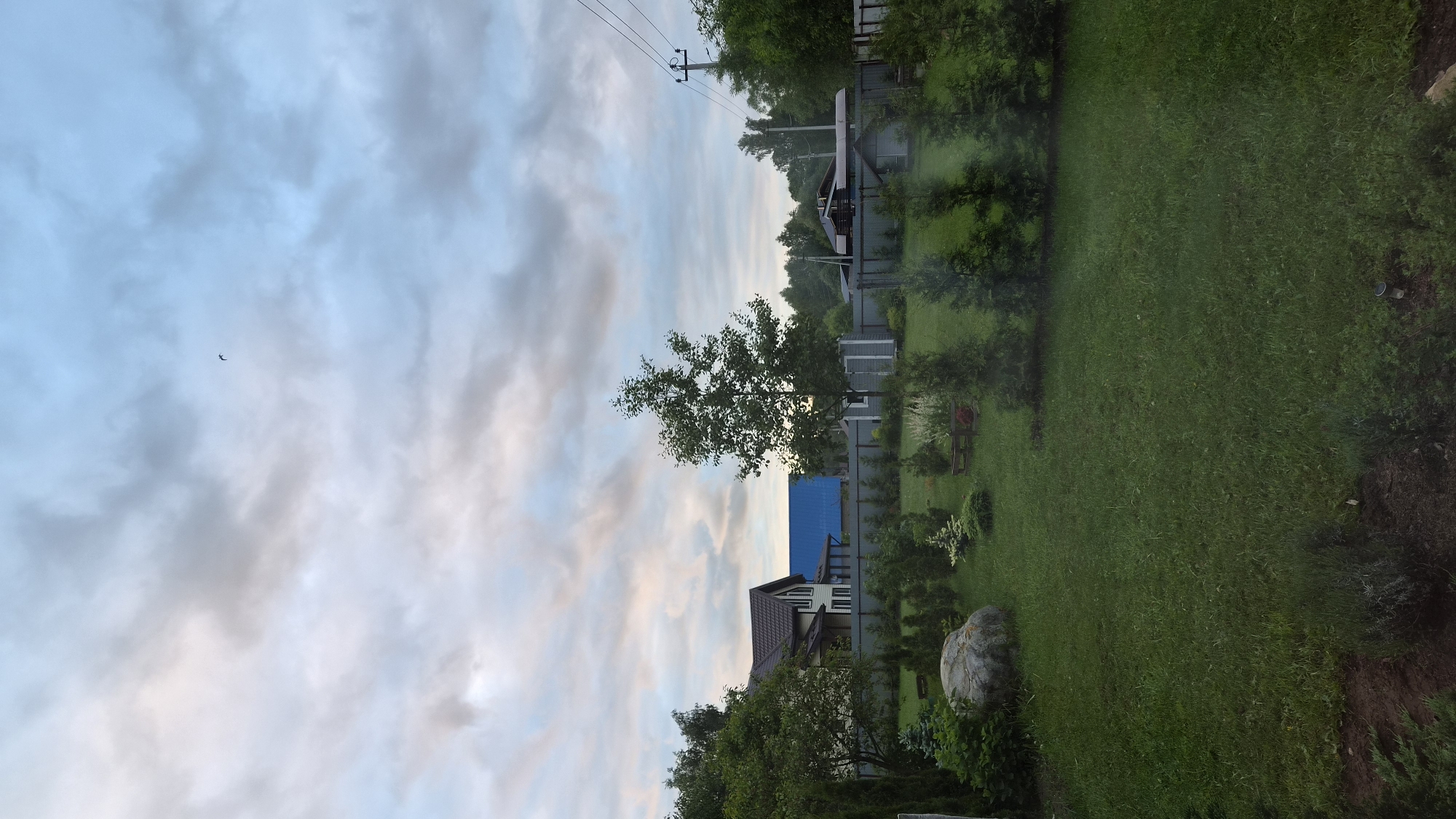 The image shows a view of a yard in Timokhovo, Russia. The sky is cloudy with patches of blue and a small object that may be a bird is visible. A house with a brown roof and a blue structure behind it is partially visible beyond a fence. A large green lawn covers a significant portion of the scene. A large rock is near the bottom of the frame. Trees and various vegetation line the perimeter of the yard. Overhead power lines are present in the distance.