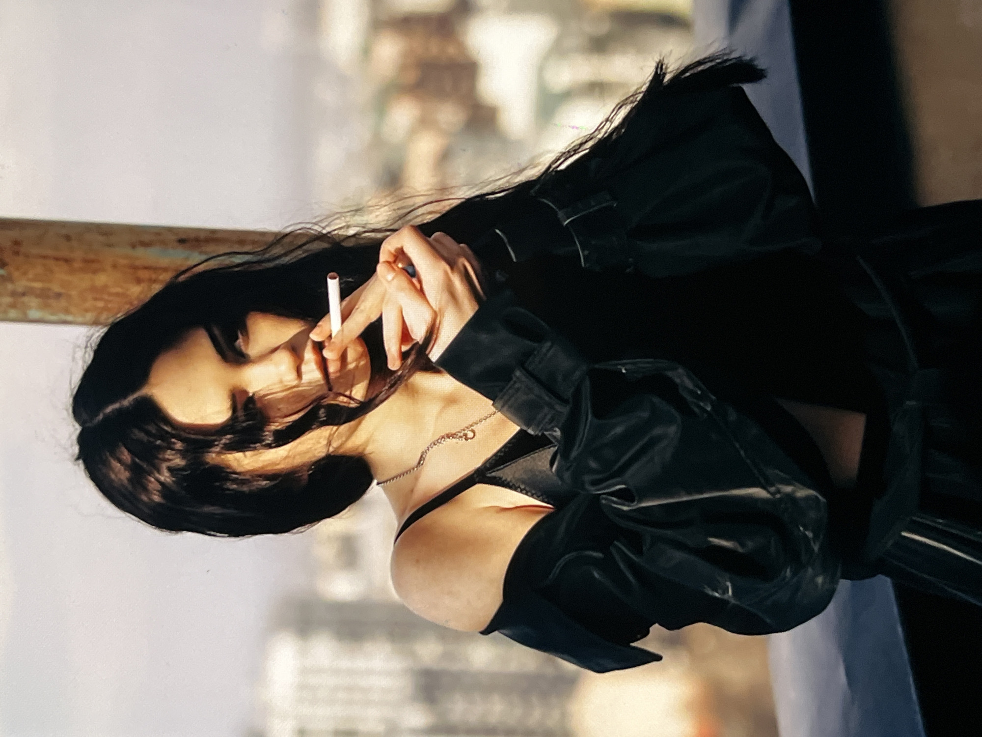A young woman with long, dark hair stands outdoors in an urban environment. She is holding a cigarette between her fingers and appears to be lighting it or has just lit it. She is wearing a black outfit, including a bra-like top and a black jacket that is draped off her shoulders. She has a necklace on. The background suggests a cityscape with buildings, and there is a metal pole to the right of the woman. The lighting is soft, possibly from the sun.