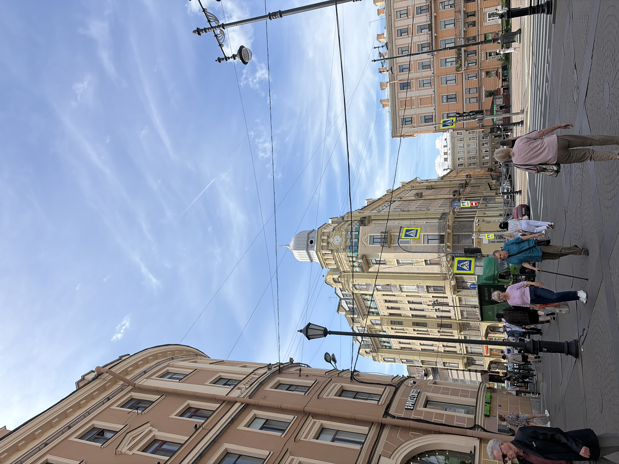 In Saint Petersburg, Russia, on a clear, sunny day, a street scene unfolds with pedestrians walking along a wide, paved sidewalk lined with multistory buildings. A prominent curved, light brown building with multiple windows and classical architectural details dominates the left side of the frame. Across the street, a yellow building with a distinctive dome-like structure, adorned with a clock face, stands out. Another peach-colored building with numerous windows is visible further down the street. Several streetlights, some with ornate designs, punctuate the urban landscape, and a network of overhead wires crisscrosses the sky.
A diverse group of people are captured in various stages of movement. In the foreground, a man in a dark jacket and sunglasses walks from left to right, while a woman in a pink top and dark pants walks away from the camera, leaning on a walking stick. Another woman in a blue shirt and brown pants is walking towards the background. Further back, several other individuals are seen walking or standing, including a woman in a light pink top and jeans, and a man in a turquoise shirt. A green truck is parked on the street. Pedestrian crossings are marked on the road. The overall impression is a bustling urban environment under a bright blue sky with scattered white clouds.