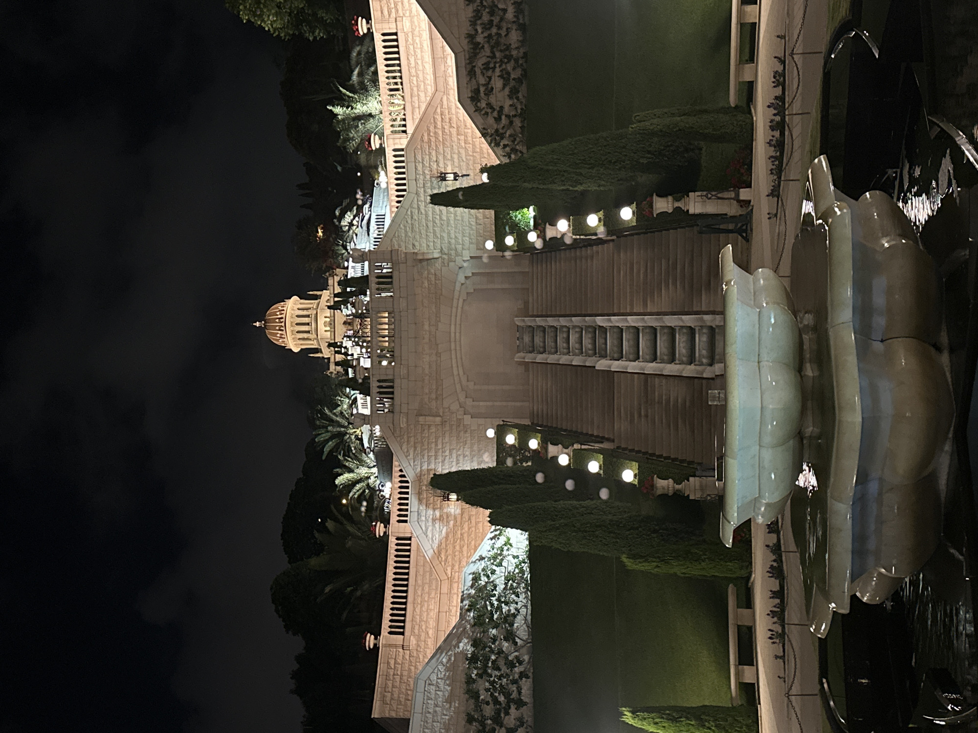 This is a nighttime, eye-level, medium shot of the Baháʼí World Centre in Haifa, Israel. In the foreground, a tiered fountain with a light green, carved basin is filled with water, reflecting the surrounding lights. Behind the fountain, a series of meticulously manicured green hedges form terraces leading up to a grand stone staircase. The stairs are flanked by tall, conical cypress trees and illuminated by spherical globe lights. Further up the slope, more stone terraces and balustrades are visible, along with palm trees and decorative planters. At the apex of the scene, the golden dome of the Shrine of the Báb is illuminated, standing out against the dark, cloudy night sky. The architecture is monumental and symmetrical, with the entire complex bathed in artificial light. No people are visible in the image.