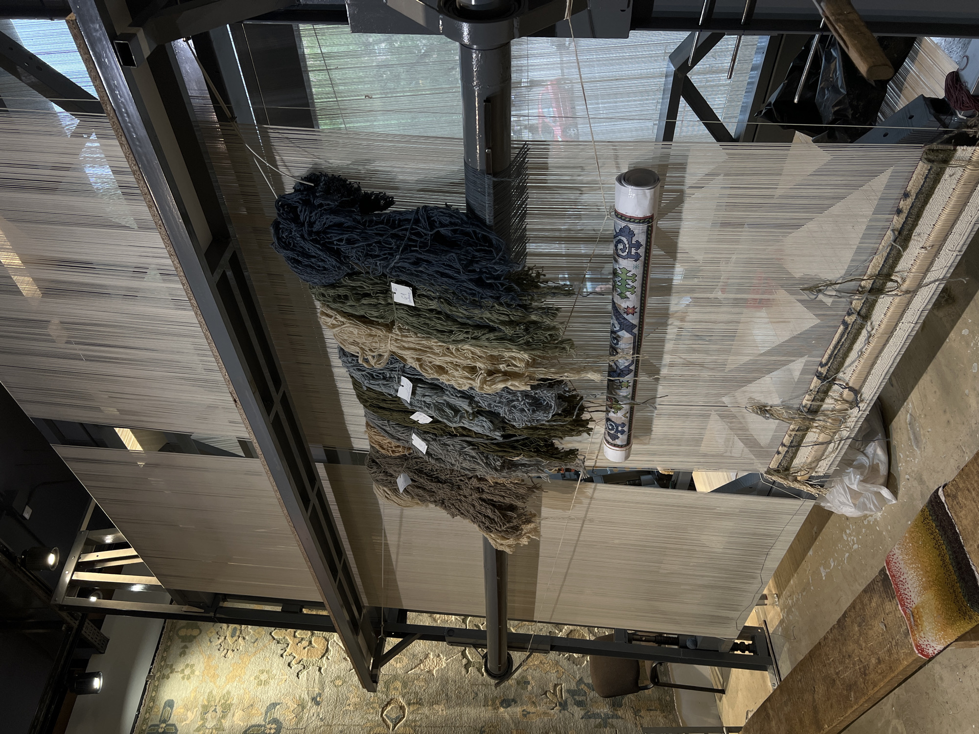 The image depicts a weaving loom in Yerevan, Armenia. The loom is made of a dark-colored metal frame. Stretched across the loom are numerous vertical white threads, forming the warp. At the top of the loom are bundles of yarn in various colors: dark blue, green, beige, and brown. These are likely the materials being used to create the weft of the carpet. A roll of patterned paper or fabric is placed near the center of the loom, possibly a guide or a sample. On the floor below the loom, a finished section of a carpet is visible. The flooring is concrete with a patterned rug beneath the machine, perhaps a showroom or demonstration of what the machine is capable of making. The light suggests it's daytime indoors.