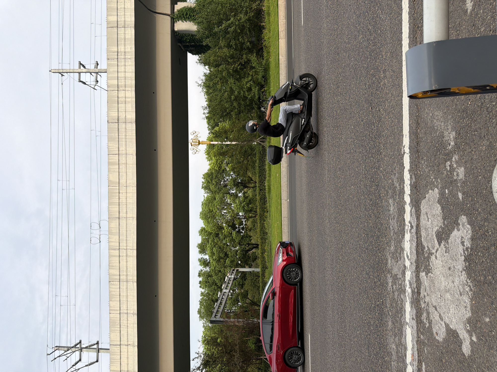 A red sedan and a dark scooter are driving on a road under an elevated highway structure. The sedan is in the left lane, and the scooter is in the right lane. The scooter is being ridden by a person wearing a black shirt, grey pants, and a black helmet. The scooter has a black storage box on the back. The elevated highway structure is concrete, and there are power lines running above it. Behind the road, there are trees and grass. The sky is overcast. The image appears to have been taken in Jiangjin District, Chongqing, China.