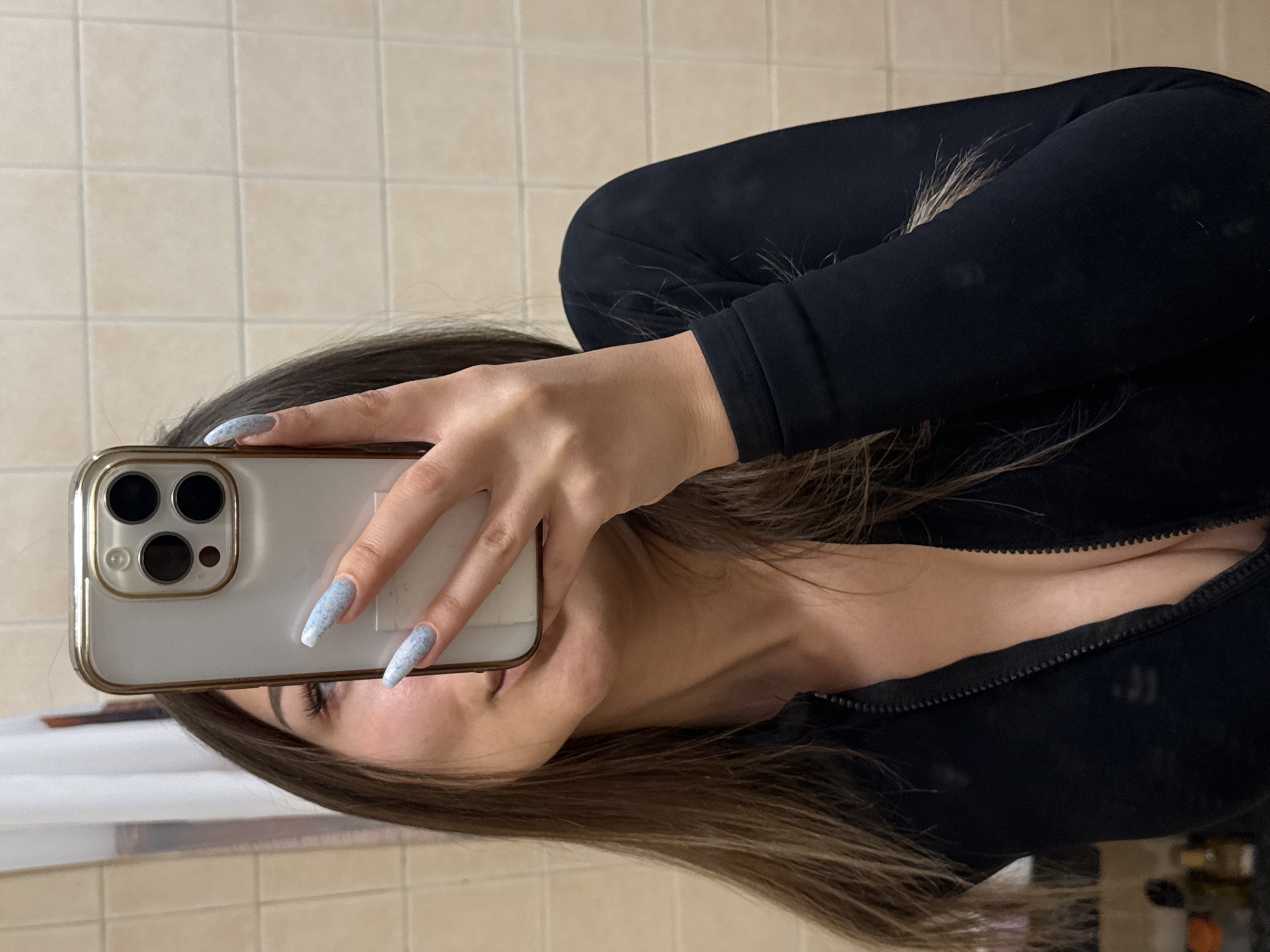A person holds an iPhone in front of their face, presumably taking a mirror selfie. The person is wearing a black long-sleeved top. Their hair is dark brown and styled. The background appears to be a bathroom with beige tiles. There is no indication that the photo was taken in Sofia, Bulgaria.