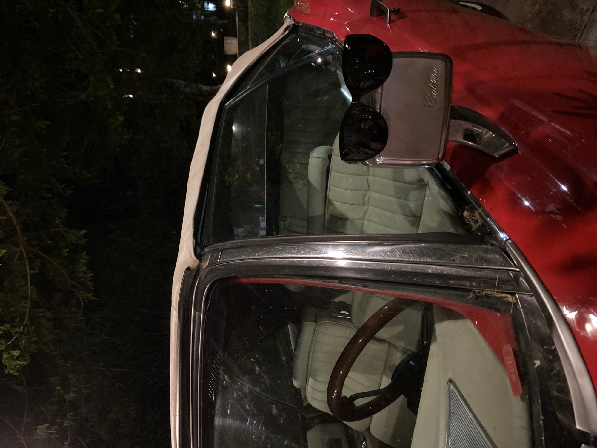 The image shows a close-up of a red Cadillac convertible parked outdoors at night. The interior is visible, with white leather seats and a wooden steering wheel. A pair of sunglasses is resting on the driver's side mirror. The Cadillac emblem is visible on the mirror casing. In the background, there are trees and what appear to be streetlights. While the exact location within Budva, Montenegro is not definitively indicated, the presence of the car and the outdoor setting suggest it could be a parking area near the city center or a residential neighborhood.