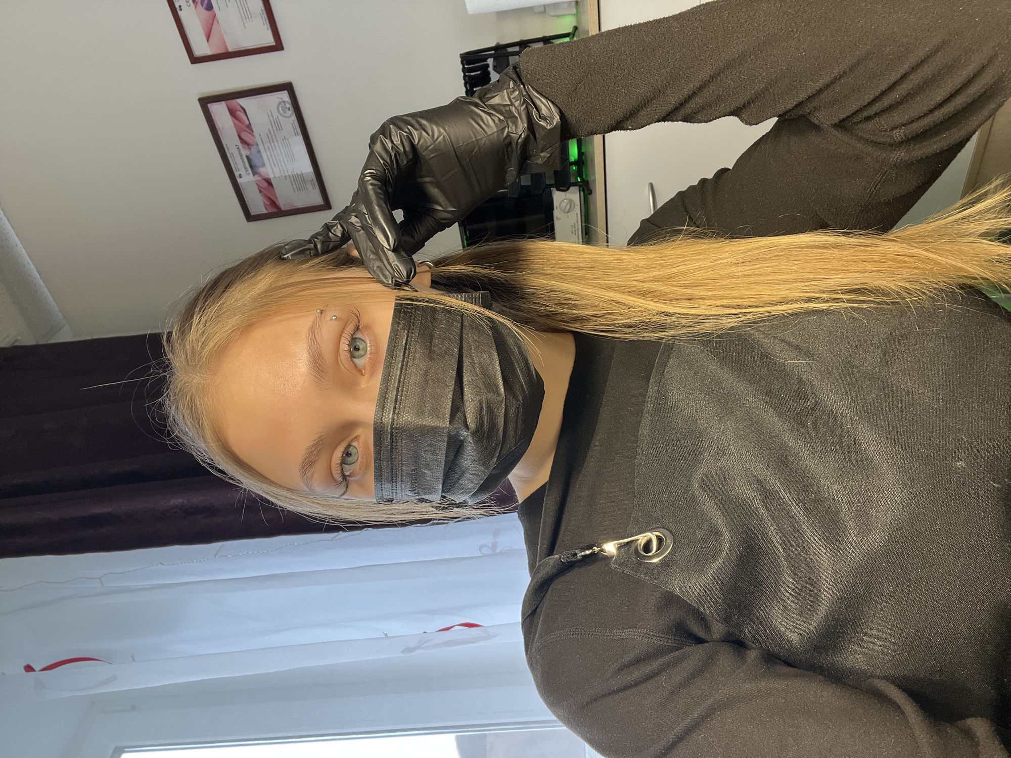 This portrait image shows a young woman in what appears to be a piercing or beauty studio in Фоки, Russia. The woman, with long, straight blonde hair, looks directly at the viewer. She is wearing a black disposable face mask, which covers her nose and mouth, and a plain black long-sleeved top that features a silver eyelet and a small black strap detail near her shoulder. Her visible left eye is a striking blue-green, and she has a silver barbell piercing on her right eyebrow.
Part of another person's arm and hand is visible above and behind the woman's head. This hand is covered by a black disposable glove and is clad in a black long-sleeved garment, suggesting a piercer or technician attending to her. The gloved hand appears to be gently adjusting her hair or preparing the area around her temple.
The background includes a white wall with two framed certificates hanging, one of which clearly displays the word "СЕРТИФИКАТ" (Certificate). A window with white blinds or curtains is also visible, allowing natural light into the room. Further in the background, a piece of professional equipment with a green light and a dial can be seen. The overall setting indicates an indoor professional environment where health and safety protocols (such as masks and gloves) are in place. The woman's visible eye suggests a neutral or calm expression. The time of day is not explicitly clear, but natural light from the window implies it is daytime.