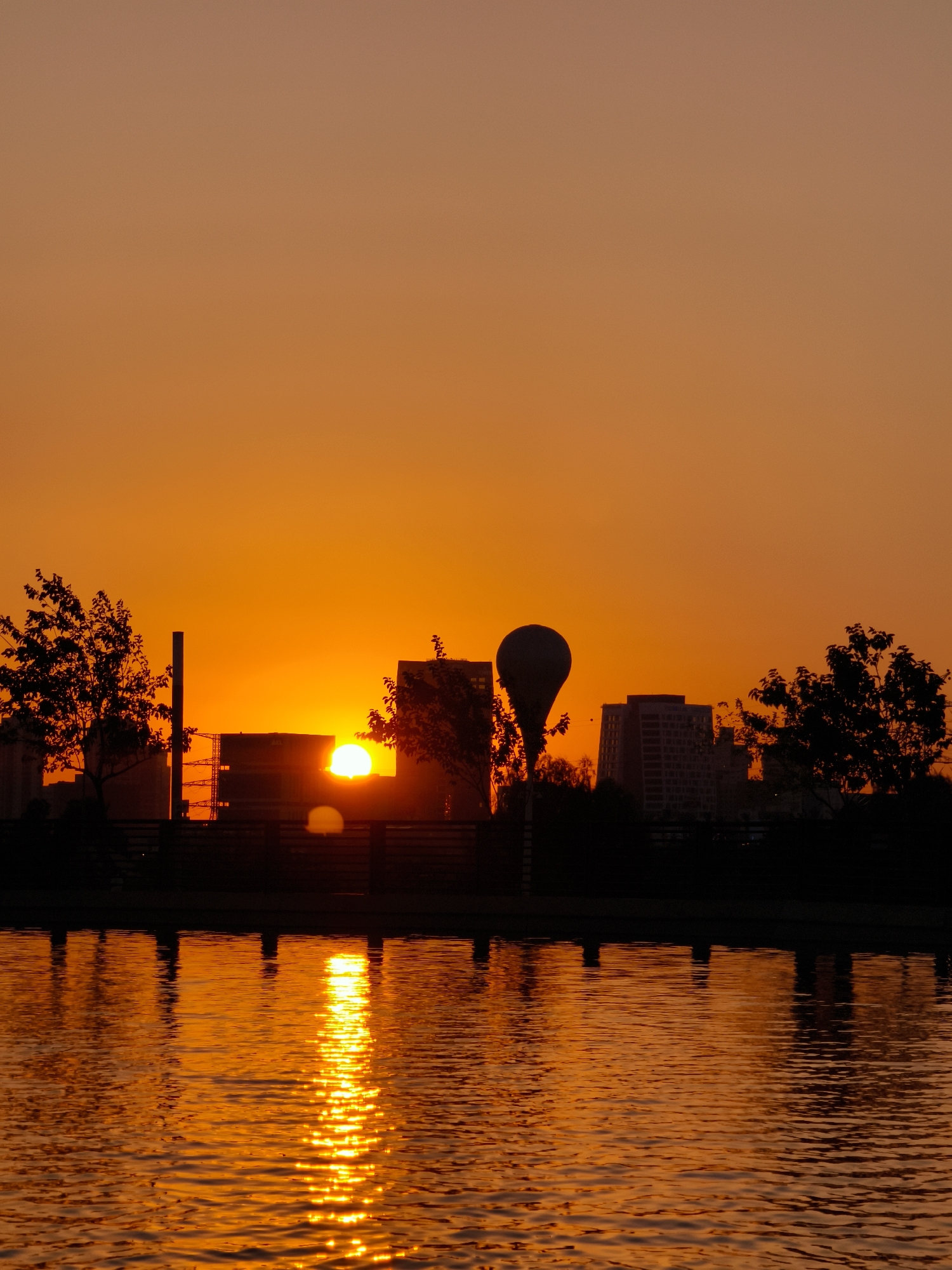 This image captures a sunset over a body of water in Be'er Sheva, Israel. The sky is a vibrant gradient of orange and yellow. Silhouetted against the bright sky are trees, buildings, and a distinctive balloon-shaped lamppost. The sun, a bright orange orb, is partially visible near the horizon, casting a shimmering reflection on the rippling water. The water's surface is a warm, golden hue, reflecting the intense colors of the sunset. The foreground features the water with its reflections, and the background shows the city skyline and vegetation in silhouette.