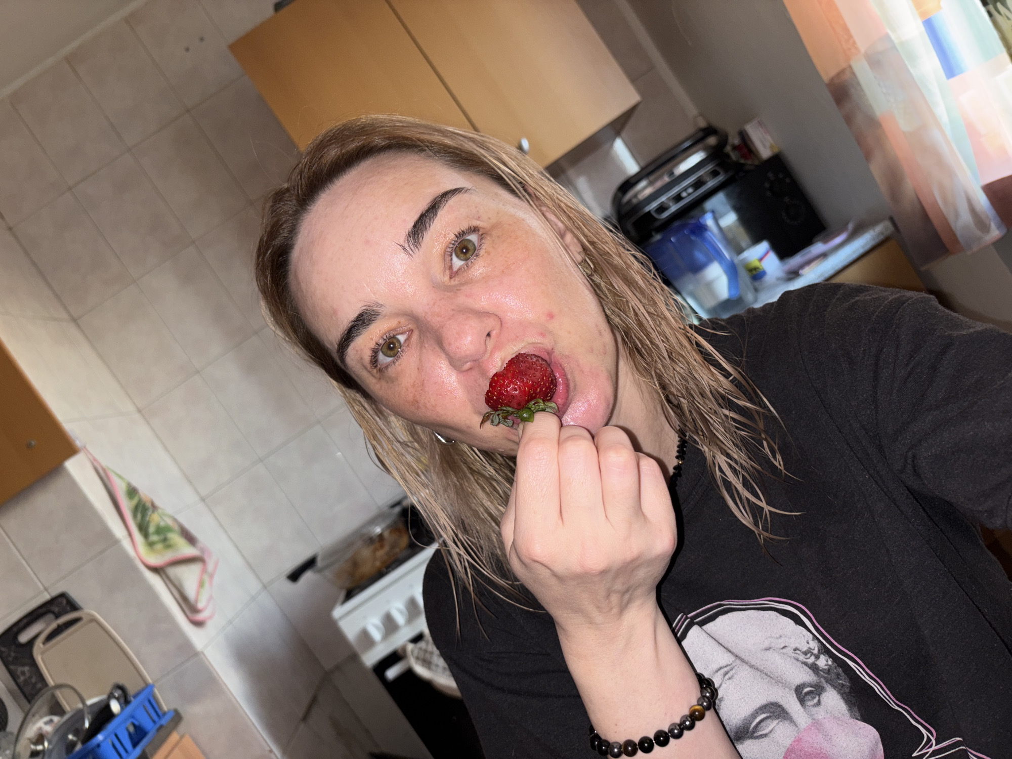 An adult woman with wet, light brown or blonde hair and light-colored eyes (green/hazel) is depicted in a close-up, upward-angled shot in what appears to be a kitchen. She has visible freckles on her face and a slight sheen, possibly from sweat or water.
The central activity shows her holding a large, red strawberry with its green stem to her open mouth, as if she is about to take a bite or is already in the process of eating it. Her eyes are wide, and she is looking directly at the camera, conveying a somewhat playful or surprised expression.
She is wearing a dark, short-sleeved t-shirt with a graphic design of a classical-style bust exhaling a pink bubblegum bubble. On her left wrist, she wears a beaded bracelet with dark and brown/golden-colored beads, some appearing metallic.
The background reveals typical kitchen elements, including light-colored wooden cabinets, tiled walls or backsplash, a stovetop or oven, and various small appliances on the counter, such as a water filter pitcher and a toaster or similar device. A multicolored, translucent curtain hangs in the upper right. A patterned kitchen towel is visible in the lower-left background, and a blue dish drying rack with dishes is partially seen. The lighting suggests an indoor setting, possibly daytime, with a mix of ambient and artificial light. There is no discernible text in the image.