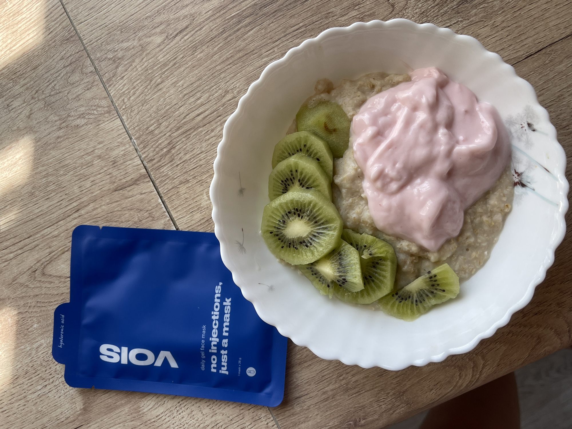 A top-down, eye-level shot shows a white, scalloped bowl filled with oatmeal, topped with slices of kiwi and a dollop of pink yogurt. Next to the bowl, on a light brown wooden surface, lies a blue packet for a "VOIS daily gel face mask" that contains "hyaluronic acid" and promises "no injections, just a mask." The lighting suggests it is daytime, with a strong shadow cast from the top left. The scene appears to be a breakfast setting, possibly in a home or hotel room, as indicated by the wooden surface which resembles flooring or a tabletop. The specific location of Мосметаллоконструкция Онт, Russia, cannot be determined from the visual information present in the image.