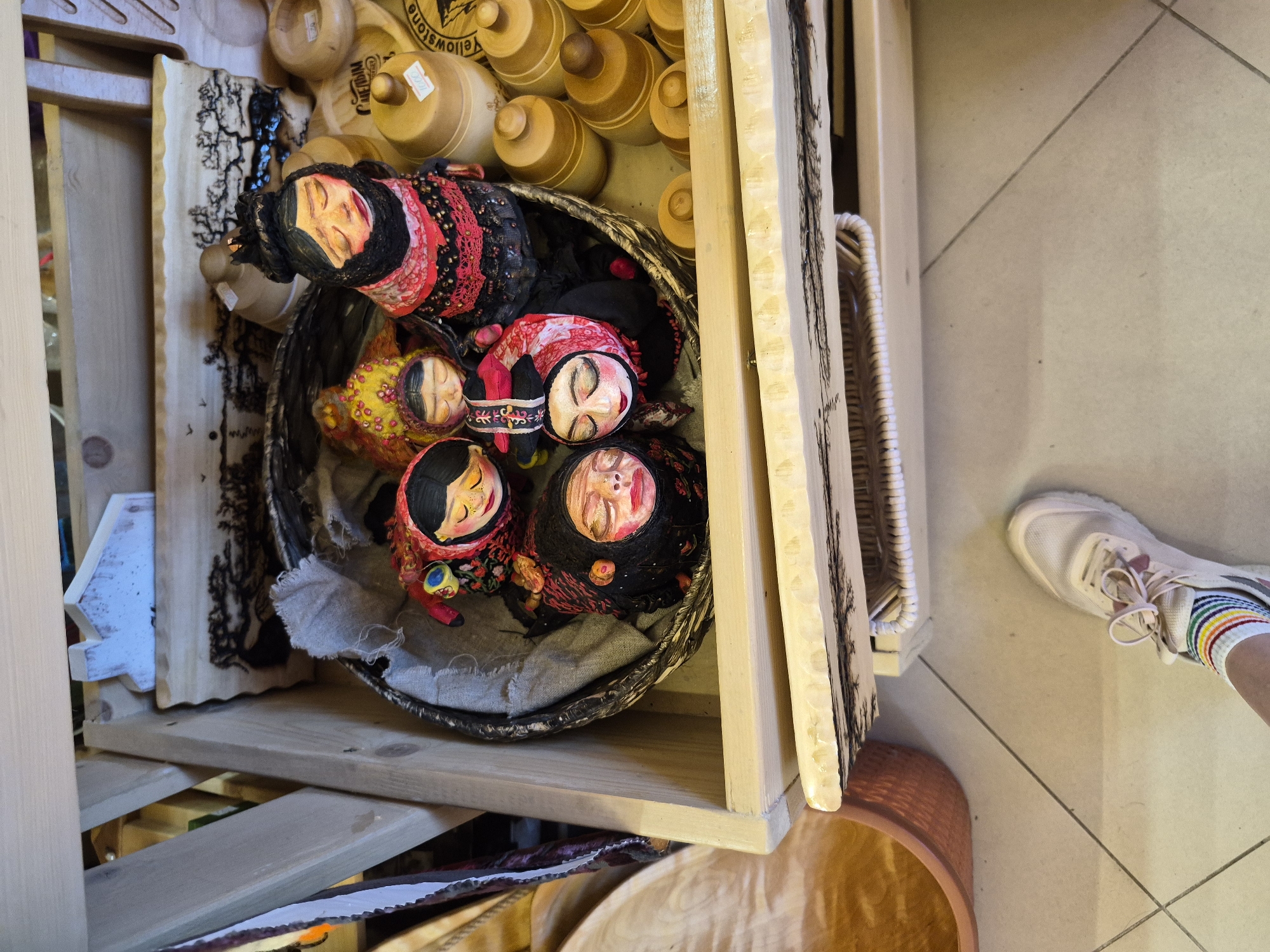The image depicts a close-up view of a wooden shelf unit filled with various handcrafted items. In the upper section of the shelf, a woven basket contains several Matryoshka dolls. These dolls are adorned with traditional attire, featuring floral patterns and scarves. Some dolls have painted faces with closed eyes and rosy cheeks, while others are partially obscured. To the right of the basket, several wooden containers with lids are stacked, some with visible labels. Below the basket, a decorative wooden plaque with a carved silhouette of trees is visible. Further down, another shelf holds a woven basket. The overall environment appears to be a shop or market stall. In the foreground, the foot of a person wearing a white sneaker and striped socks is visible, suggesting the perspective of someone browsing the merchandise. The floor is tiled in a beige color with dark grout lines. The scene is lit with natural or soft artificial light, creating a warm and inviting atmosphere. This scene is likely from the city of Krasnyy Klyuch, Russia, given the presence of traditional Russian dolls.