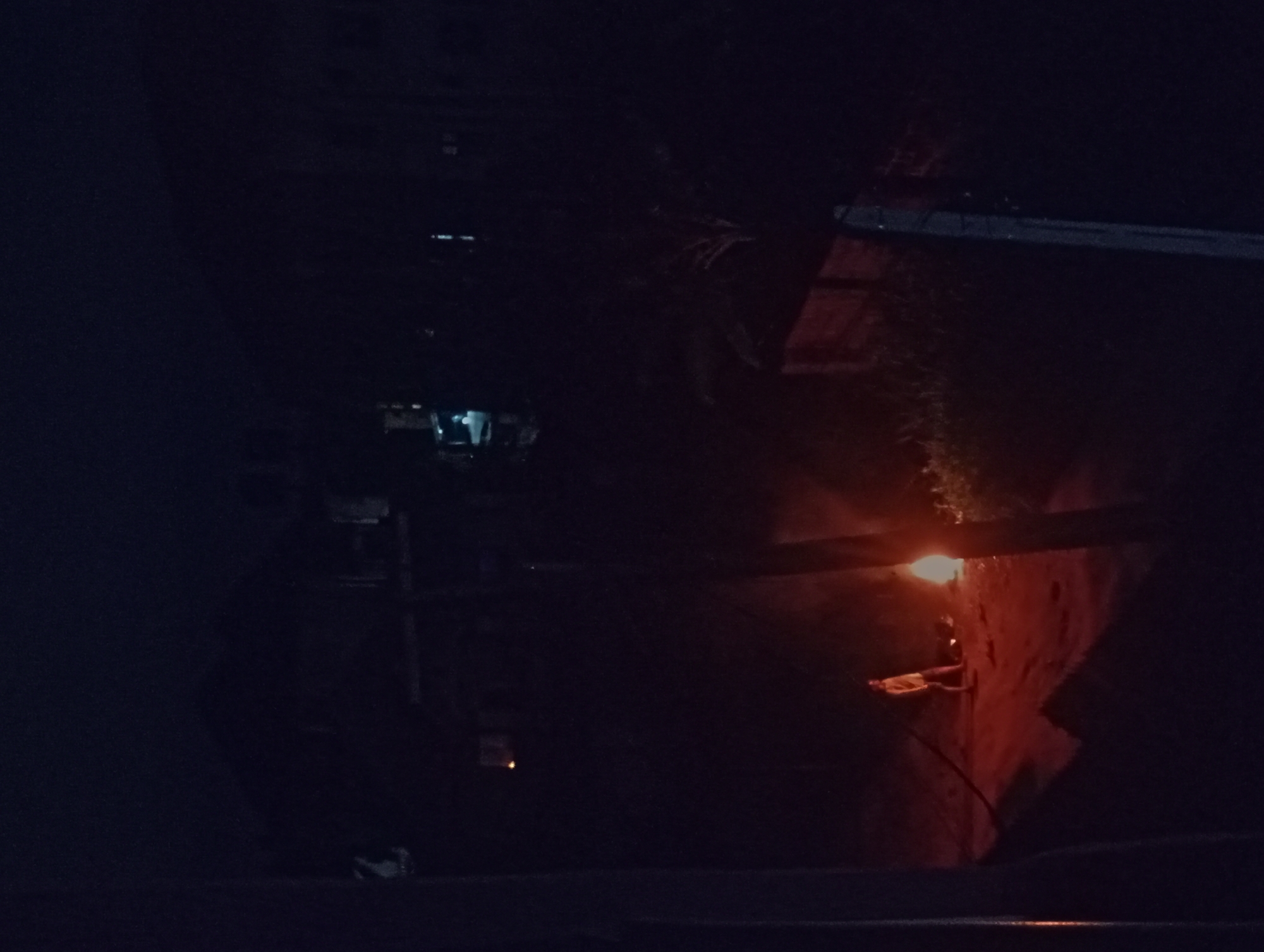The image depicts a dimly lit alleyway at night in an unknown city. The alley walls are partially illuminated by a single warm-toned light source, possibly a bulb, creating strong shadows. In the distance, there are hints of building structures. The general environment appears to be urban and potentially derelict due to the poor lighting and visible wall textures. No people are clearly visible in the frame.