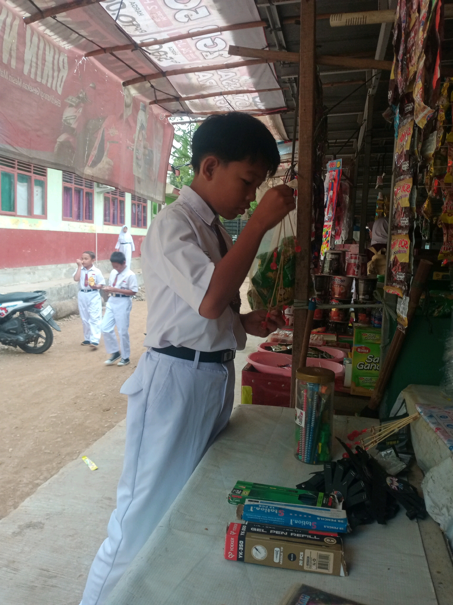 In Bandar Lampung, Indonesia, a young boy in a white school uniform, complete with a black belt, stands at a table outside a shop. He is focused on a collection of colorful string toys, his hands manipulating the strings. Behind him, the shop is stocked with various items, including packaged snacks and stationery. The shop itself is covered by a rustic awning made of corrugated metal and wood, and is partially obscured by hanging banners and plastic sheeting. In the background, other students in similar uniforms walk by, and a motorcycle is parked nearby. The scene appears to be a typical day in front of a local shop, possibly near a school.