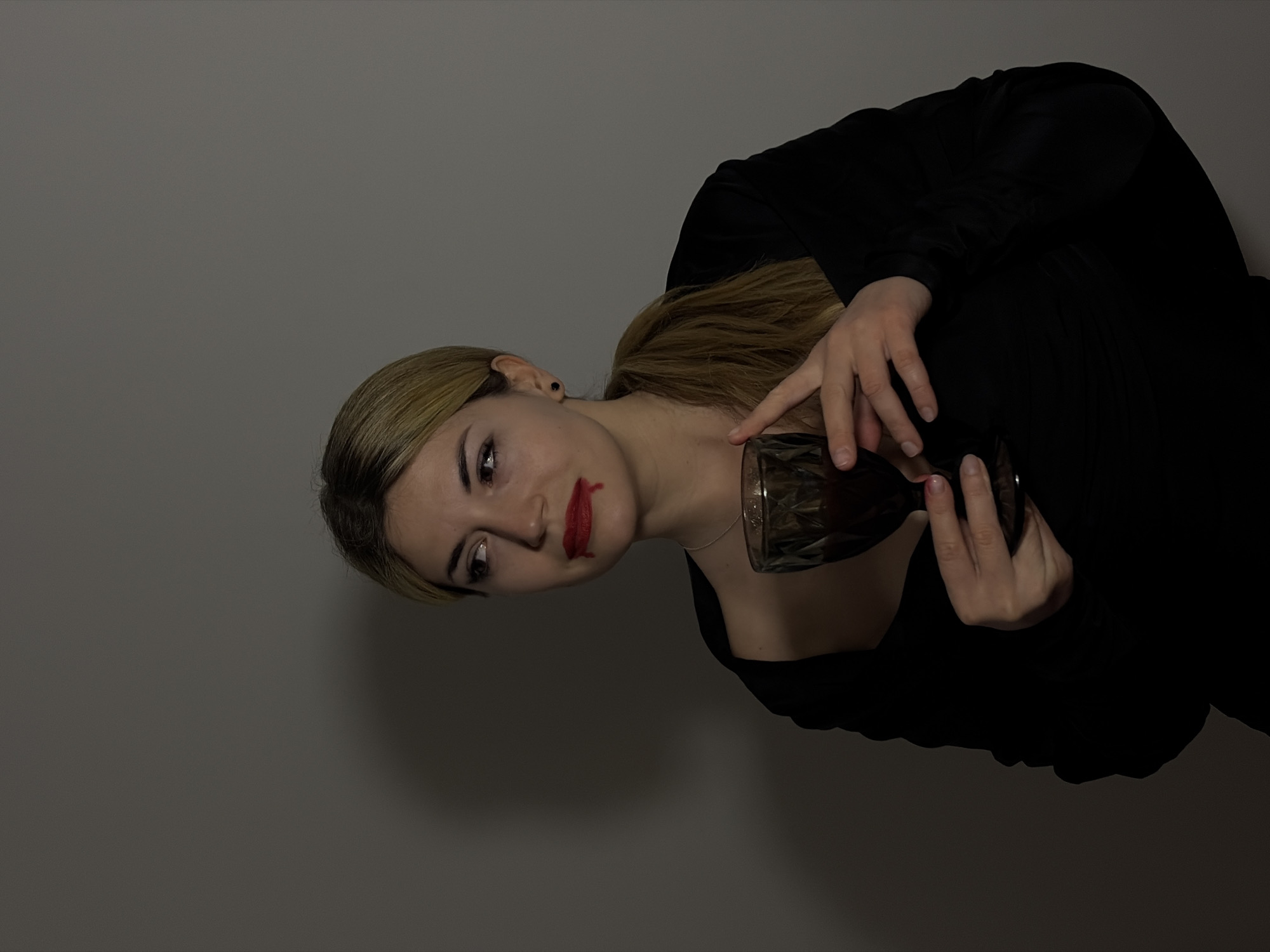 A woman with blonde hair pulled back in a ponytail is angled in the frame against a gray wall. She wears a black top. Red lipstick is smudged on her face. She is holding a glass. There is no evidence to suggest this image was taken in Budva, Montenegro.
