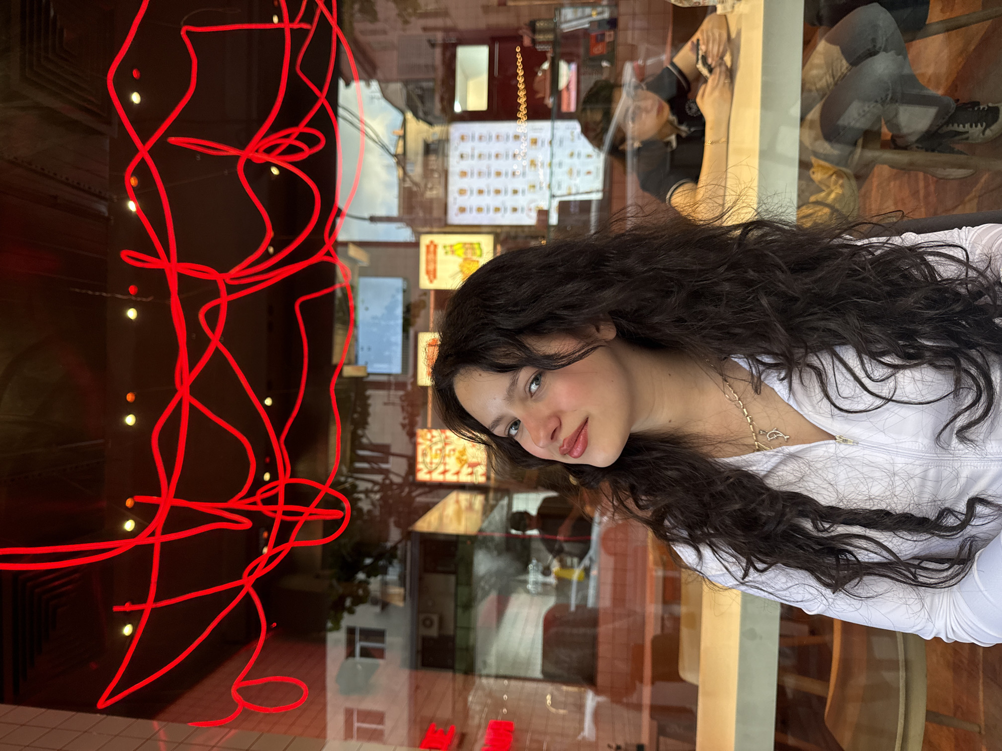 A young woman stands indoors against a large window displaying both a red neon light fixture and street reflections, notably featuring buildings and a sign. She has dark hair and wears a white top. Reflected within the glass is a partial view of another individual seated at a table. The location is Yerevan, Armenia, as indicated by visible architectural details common to the city.