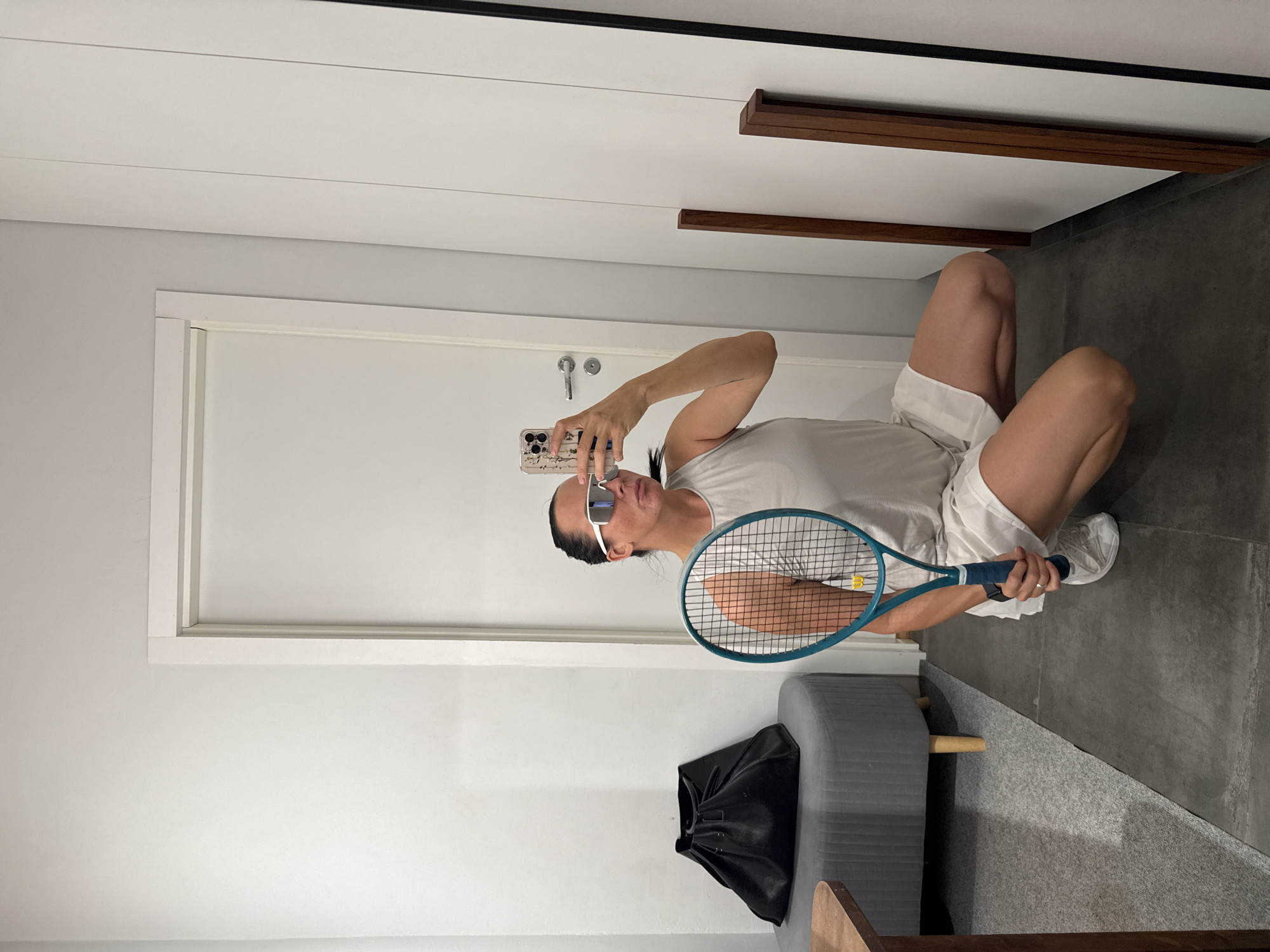 A woman wearing a light-colored sleeveless top, white shorts, white sneakers, and white sunglasses is crouching in what appears to be a dressing room or a space with closet doors and a regular door. She is holding a teal tennis racket with both hands and taking a selfie with her phone, which is visible in her right hand. A large black bag is on a grey ottoman to her left, and a brown table is partially visible in the lower left corner. The floor is a dark grey tile, and the walls are white. The image does not provide any specific visual cues that would allow for its location to be identified as Chaikovsky, Russia.