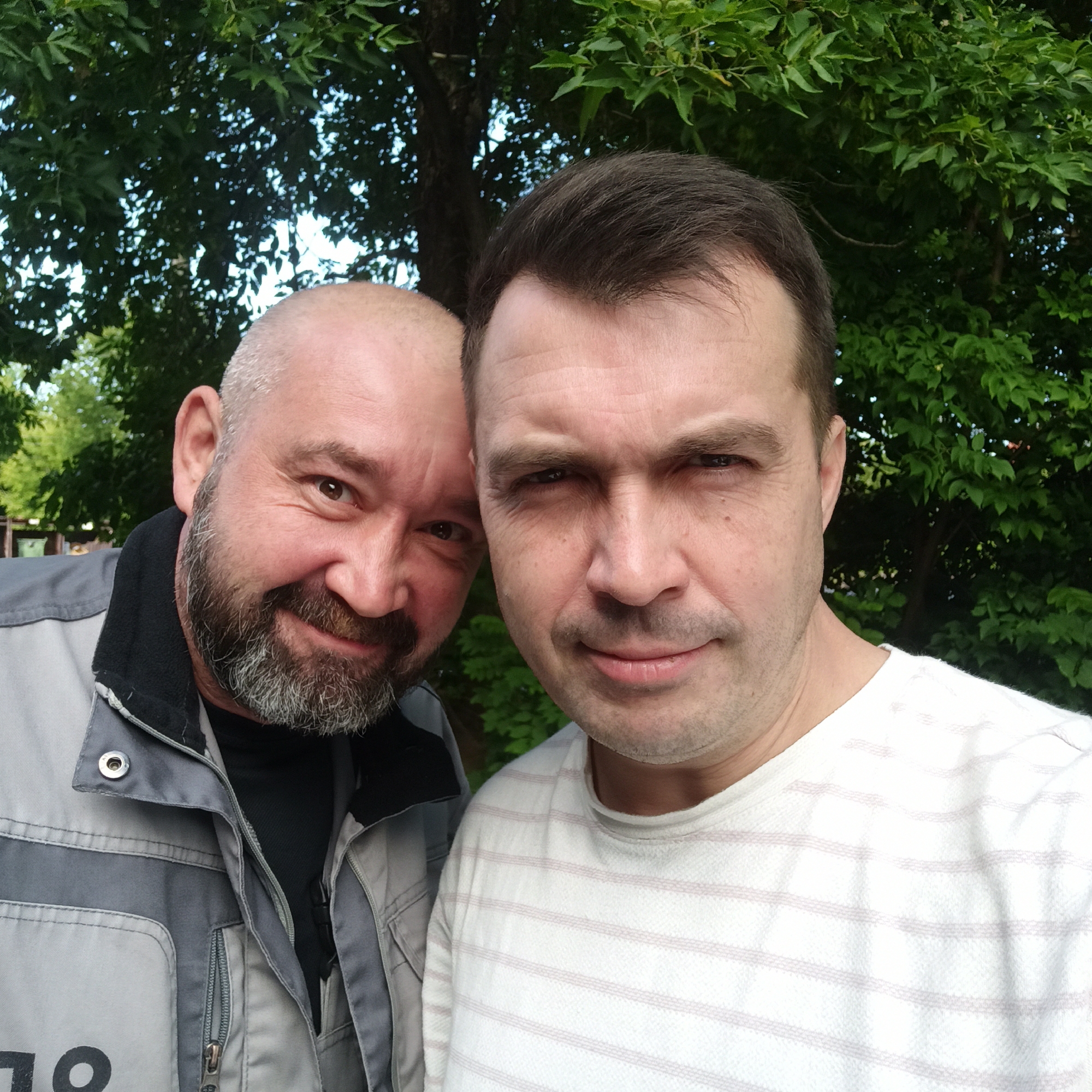 Two men, one with a bald head and beard, the other with dark hair, pose for a selfie outdoors. The man with the bald head is wearing a gray jacket with black trim and appears to be smiling. The man with dark hair is wearing a white striped shirt and looking forward. They are standing in front of a backdrop of green trees. There is no indication of the specific location in Nizhny Novgorod, Russia.