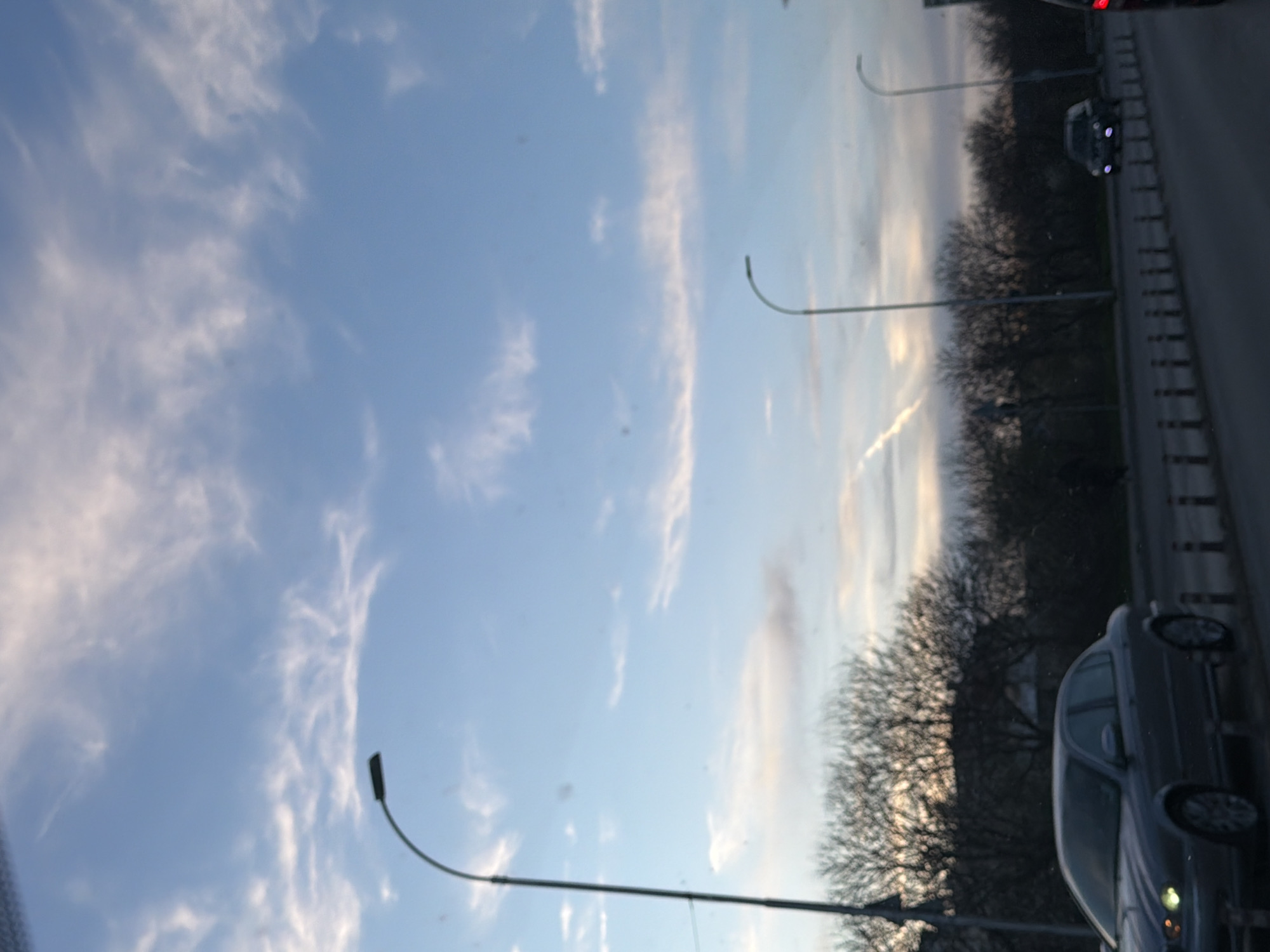 The image shows a view from a moving vehicle, likely a car, on a road in Varna, Bulgaria. The sky is visible with light blue and wispy white clouds. A row of bare trees lines the right side of the road, interspersed with streetlights. Several cars are visible on the road, including a gray car in the foreground. The road appears to be a multi-lane highway with guardrails.
