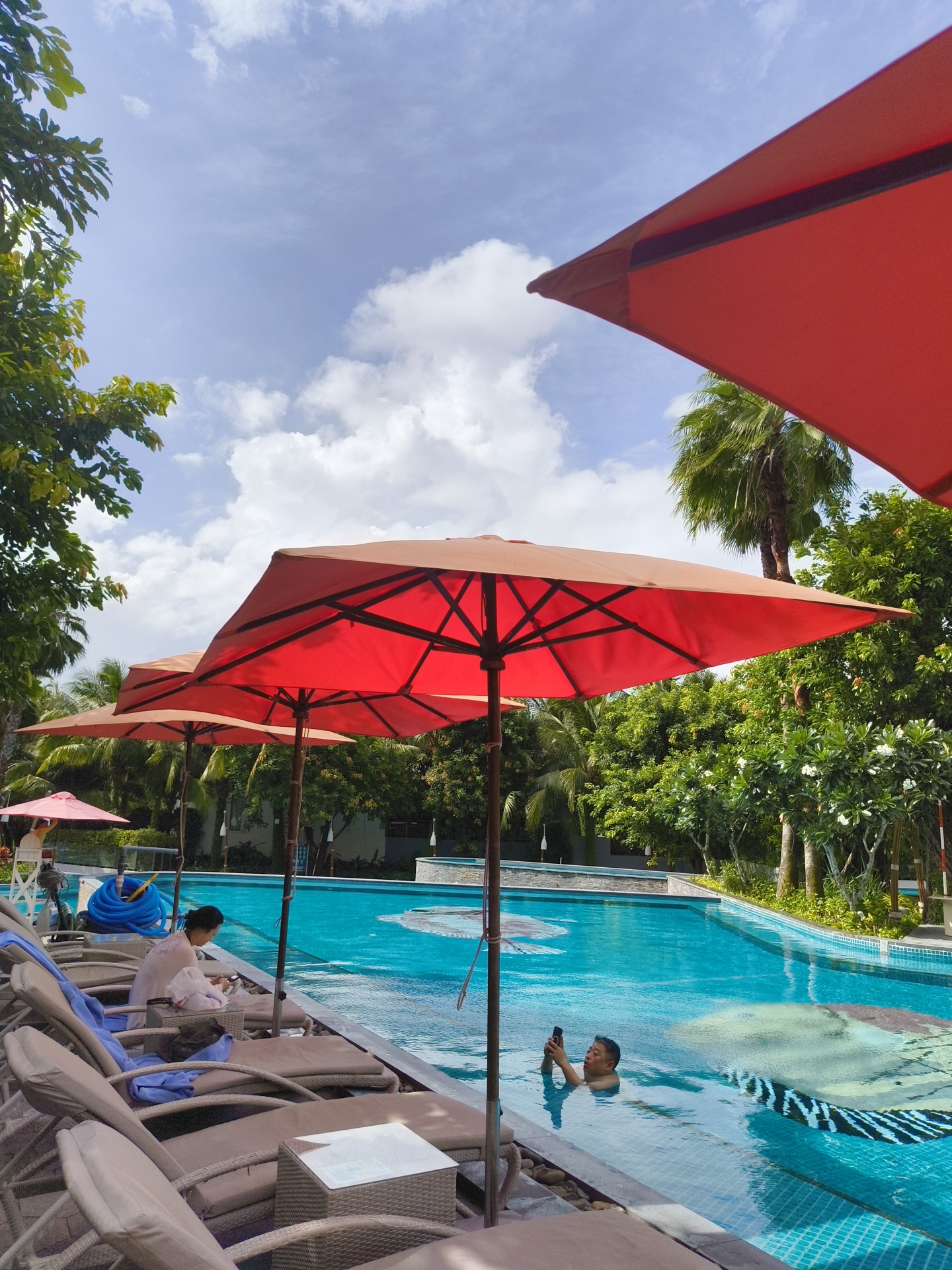 A man is in a swimming pool, holding a smartphone. He is in the water up to his chest and looking at his phone. The pool is a bright blue color and has a patterned bottom. Around the pool, there are lounge chairs and large red umbrellas. In the background, there are palm trees and lush green foliage. The sky is blue with white clouds. This appears to be a resort or hotel pool in Phú Quốc, Vietnam.